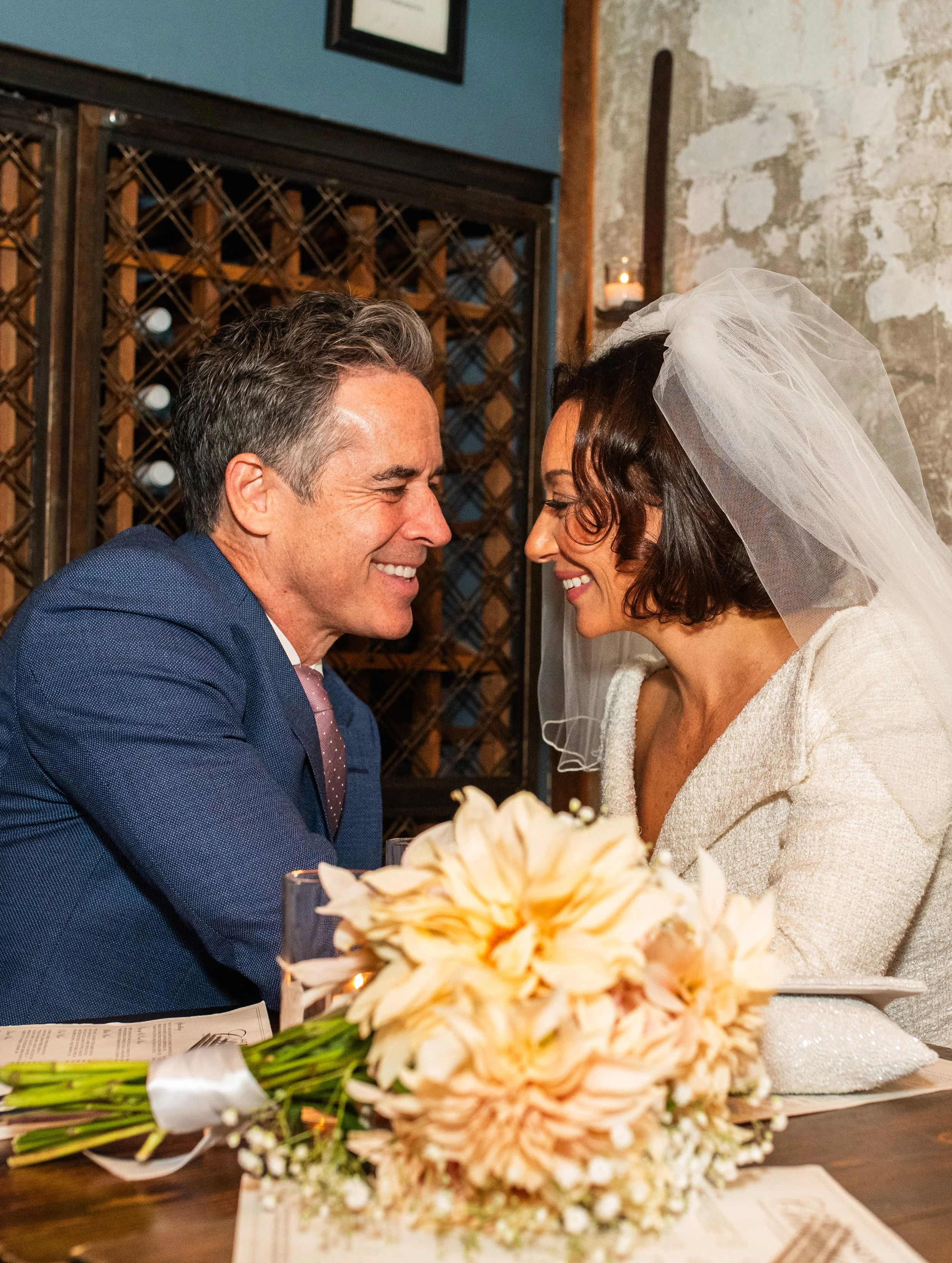 Bride and groom seated, leaning towards each other smiling with the bouquet on the table after their Cakewalk elopement ceremony.