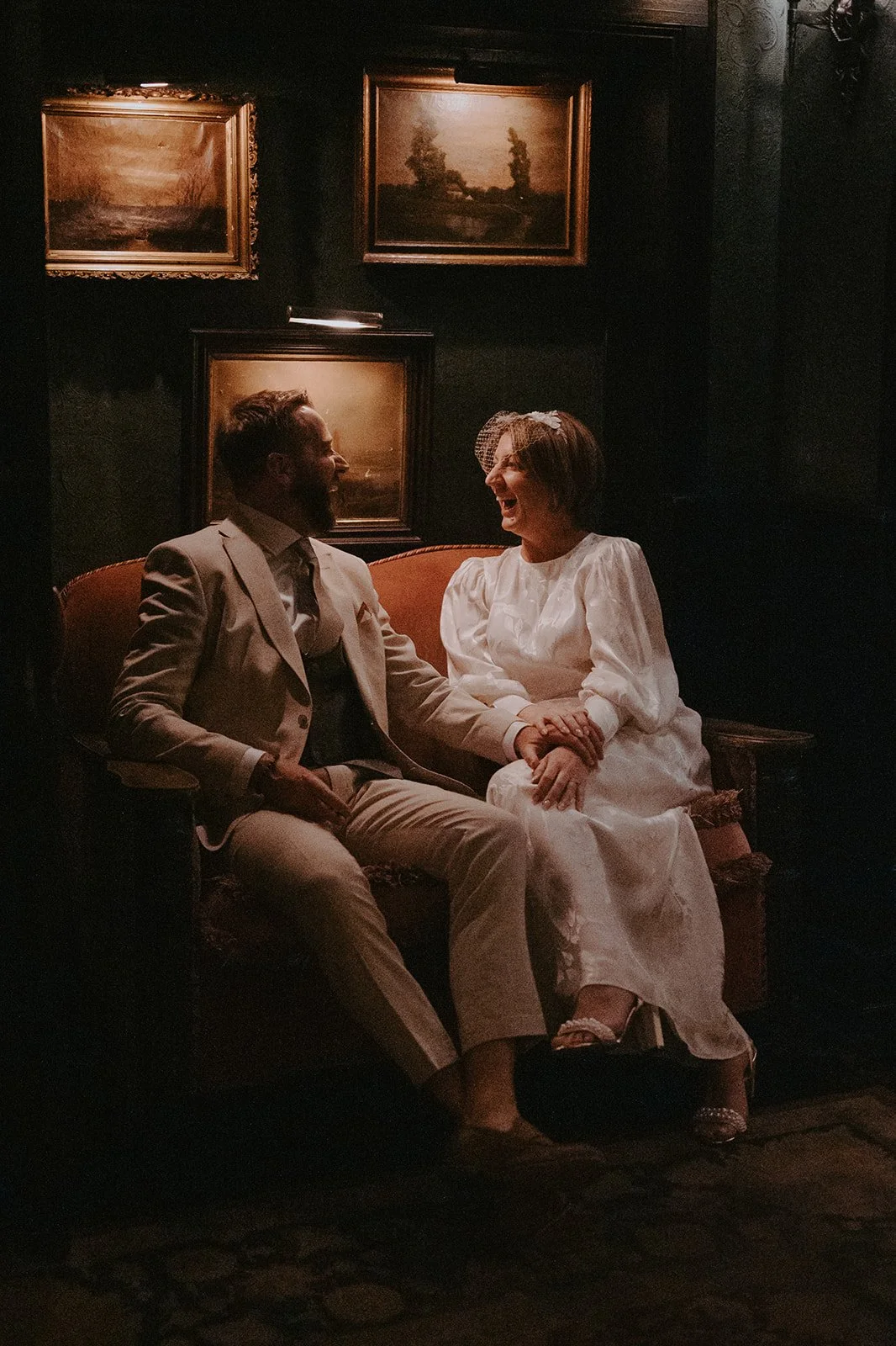 Bride and groom seated in a velvet chair in mood lighting inside a bar with paintings on the wall behind them after their Cakewalk elopement.