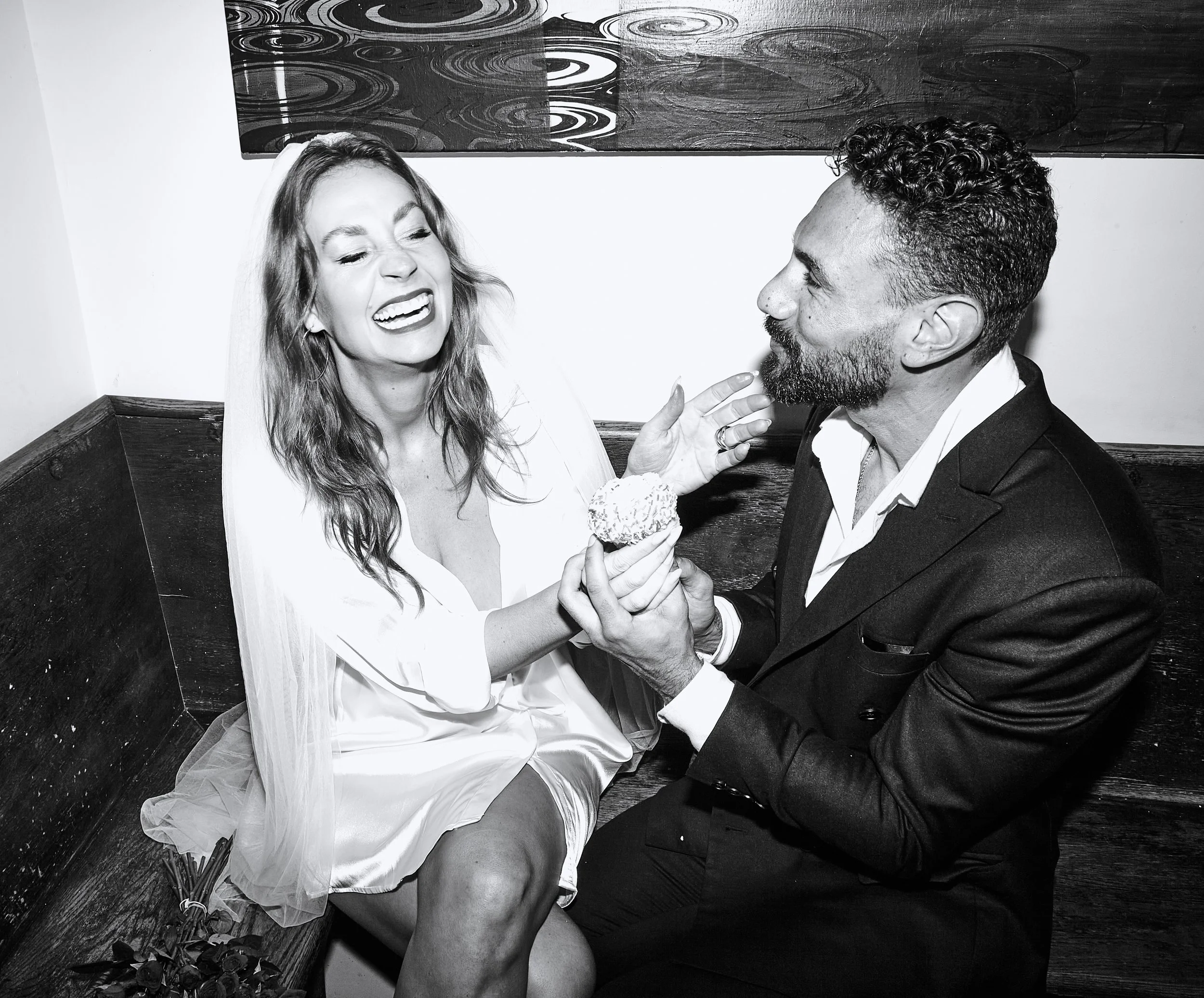 Bride and groom smiling and sharing a donut while seated on a bench after their Cakewalk wedding in Brooklyn.
