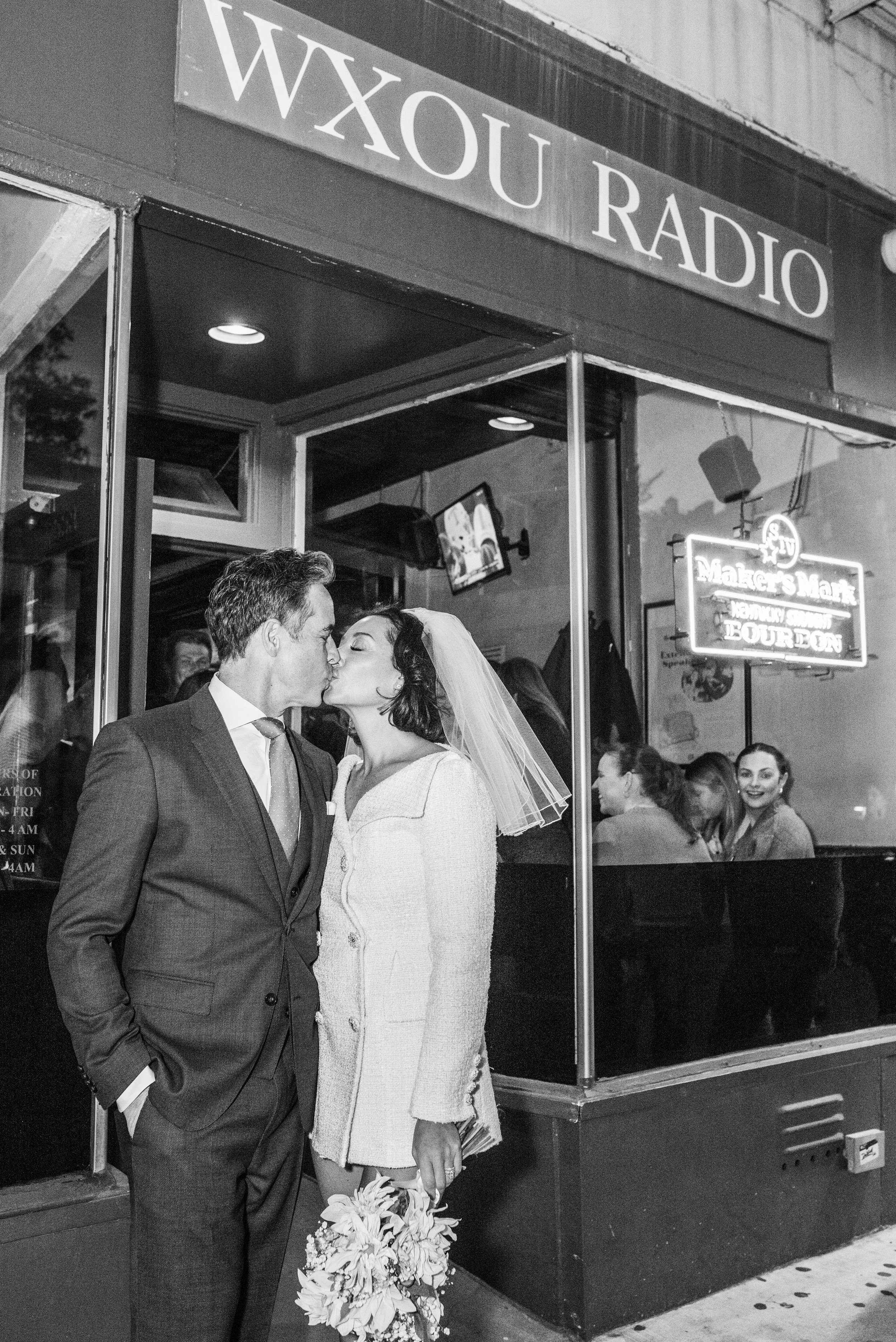 Bride and groom kissing outside of WXOU Radio where the beer sign glows and customer admire the couple. 