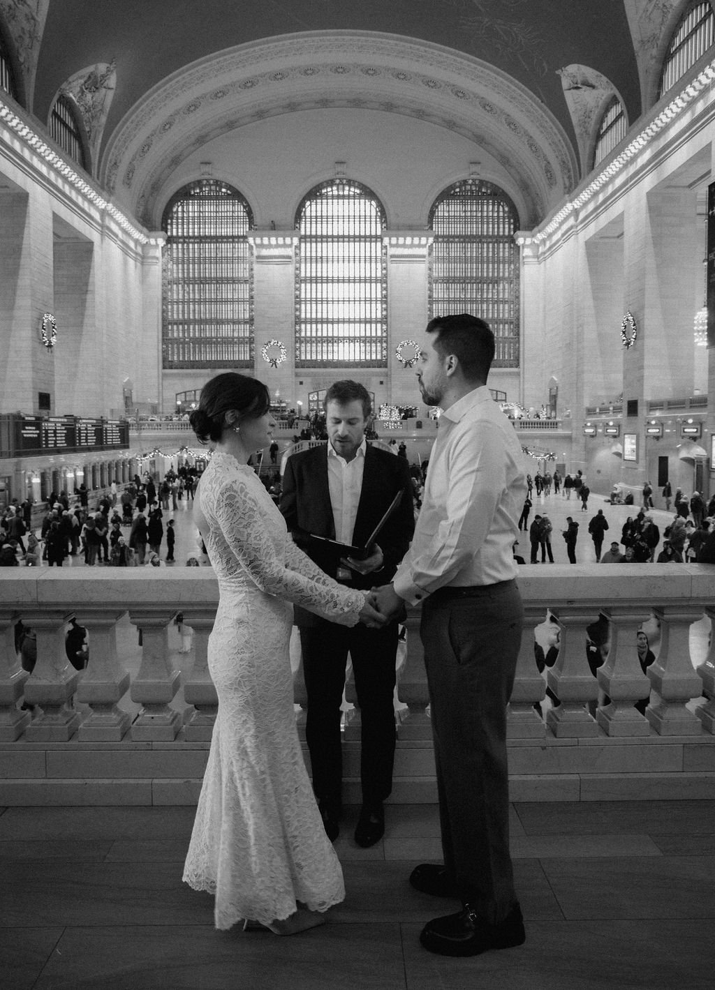 Black and white photo of a couple getting married at Grand Central Station, an officiant standing between them, holding a book, and people in the background.