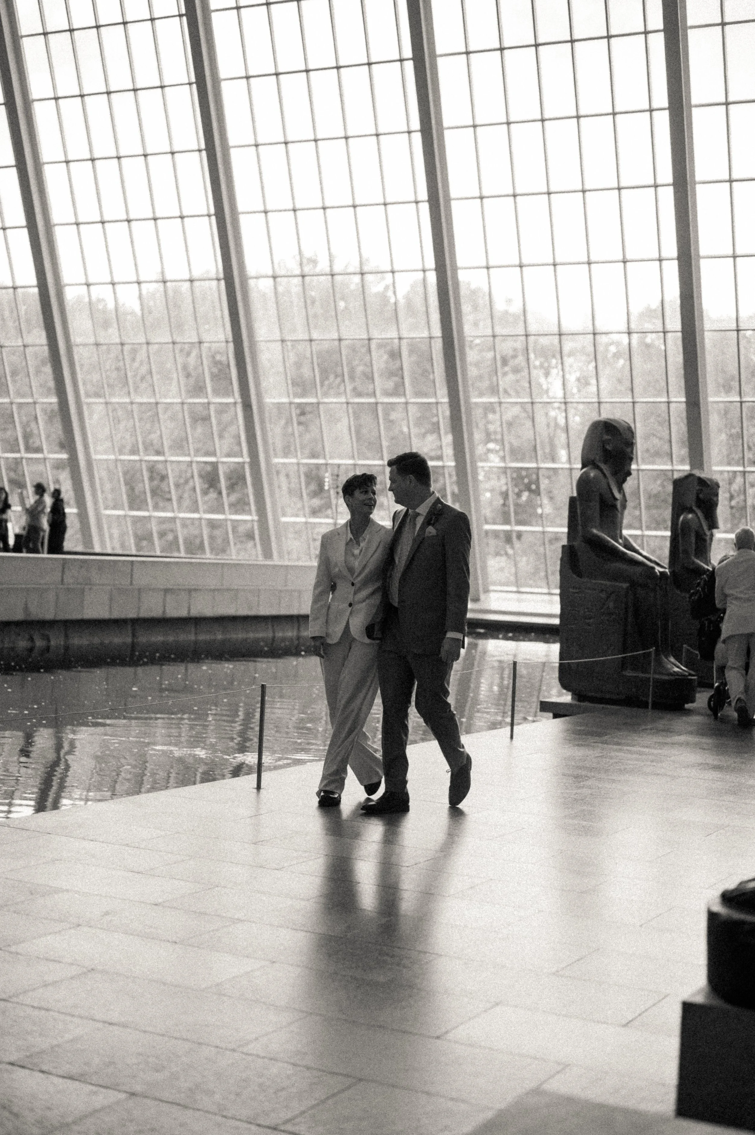 Same-sex couple walking hand-in-hand through The Met’s Temple of Dendur with statues and glass walls around them.