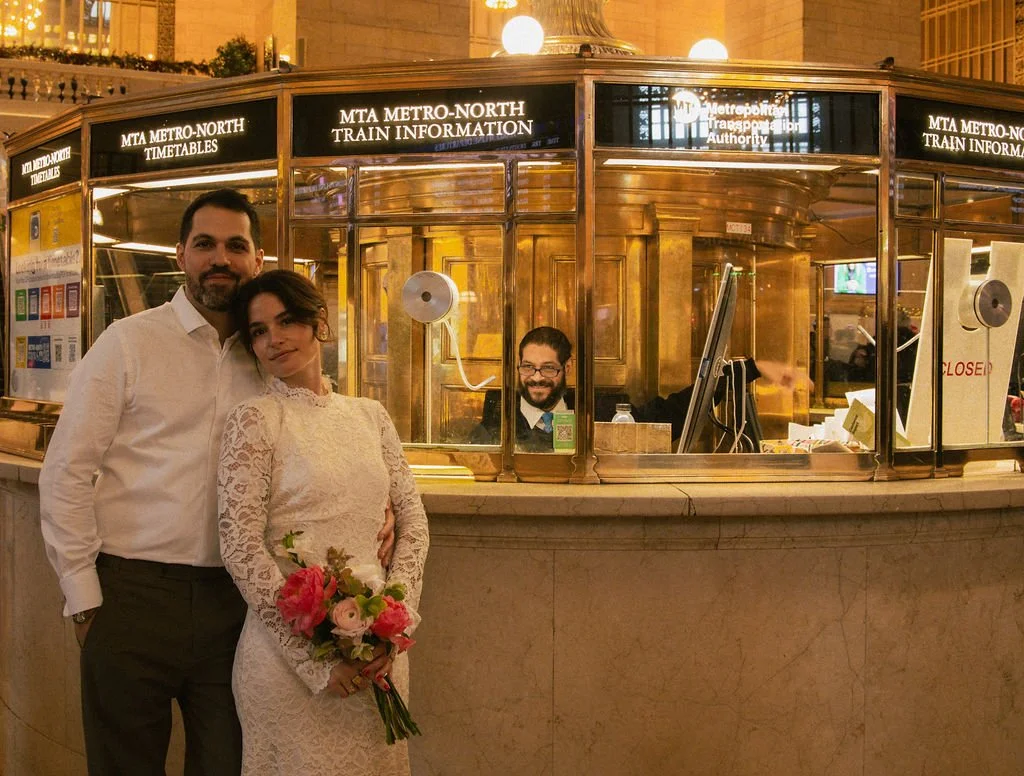 Married couple with a bouquet standing at the ticketing office at Grand Central Station after their Cakewalk elopement ceremony.