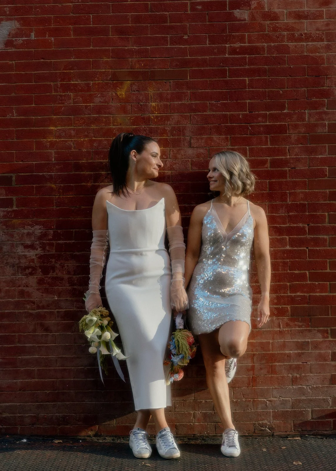 Australian lesbian brides posing in front of a brick wall in Manhattan, holding flowers and wearing tennis shoes after their Cakewalk elopement.
