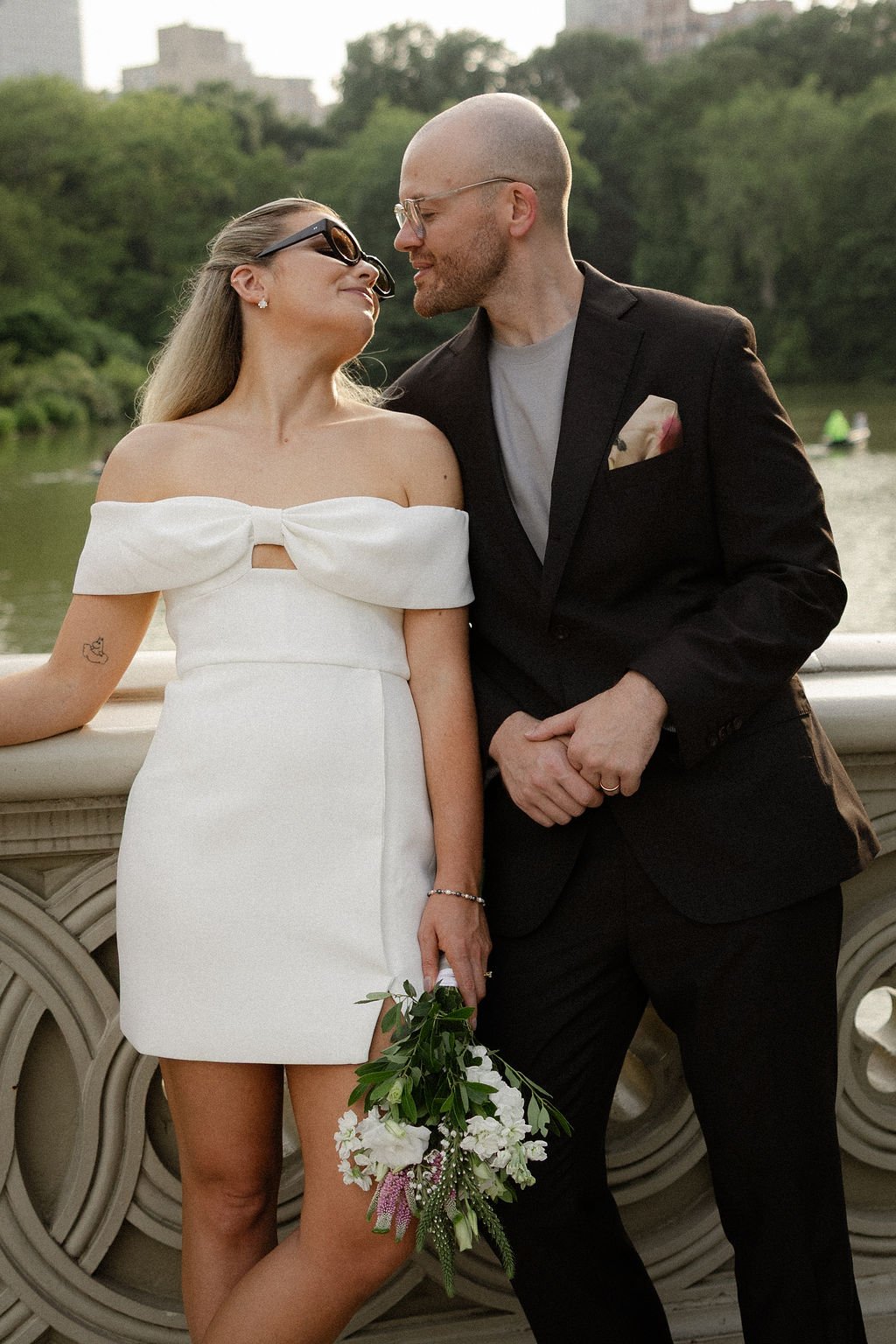 Bride and groom posing on a bridge in Central Park with greenery and water behind them.