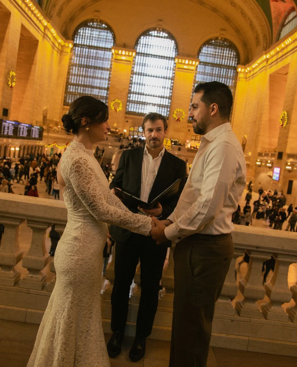 A couple getting married indoors at a grand, historic train station, with an officiant standing between them, holding a book, and people in the background.
