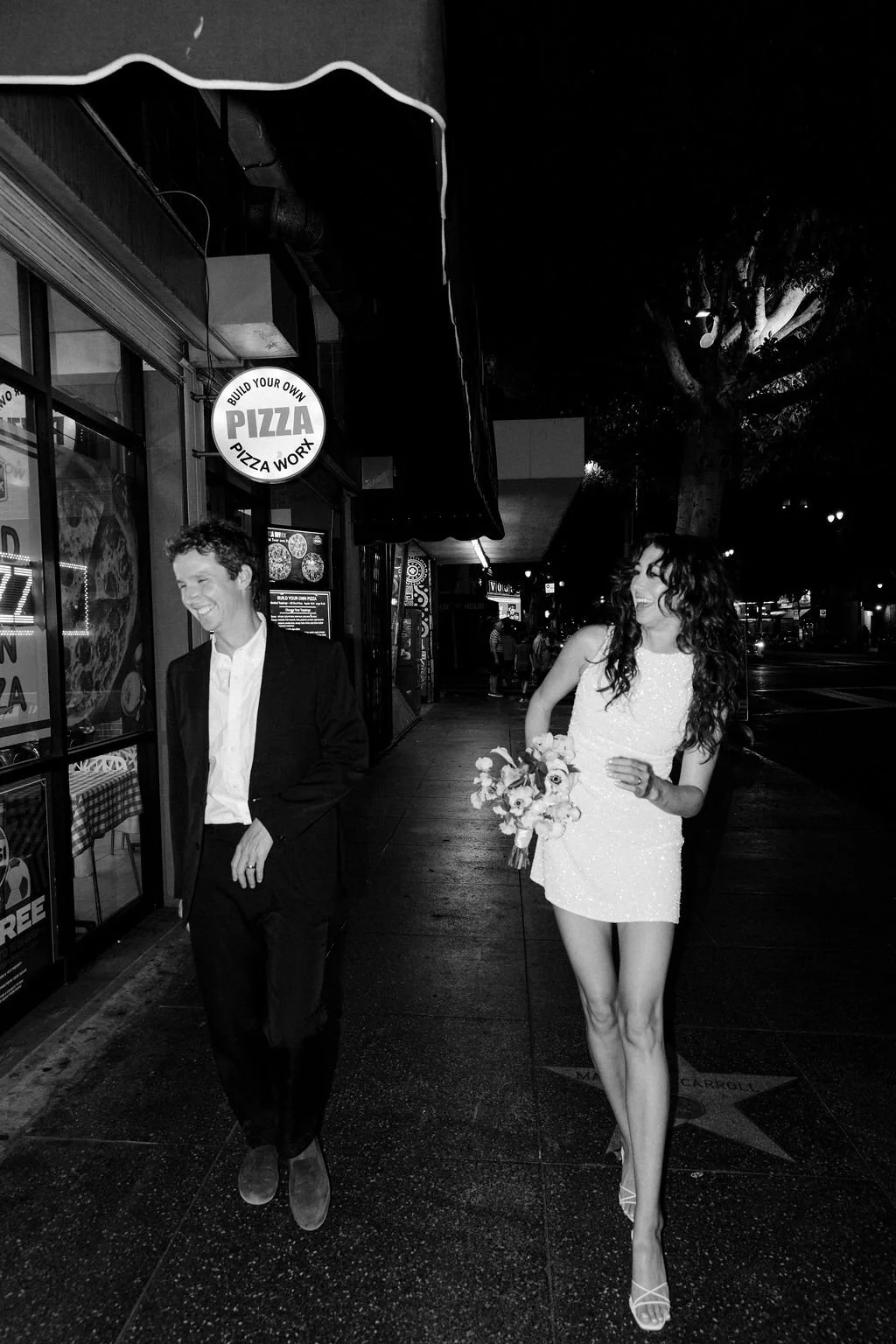 Couple walking on Hollywood Blvd in front of a pizza shop on the Walk of Fame at night in wedding attire.