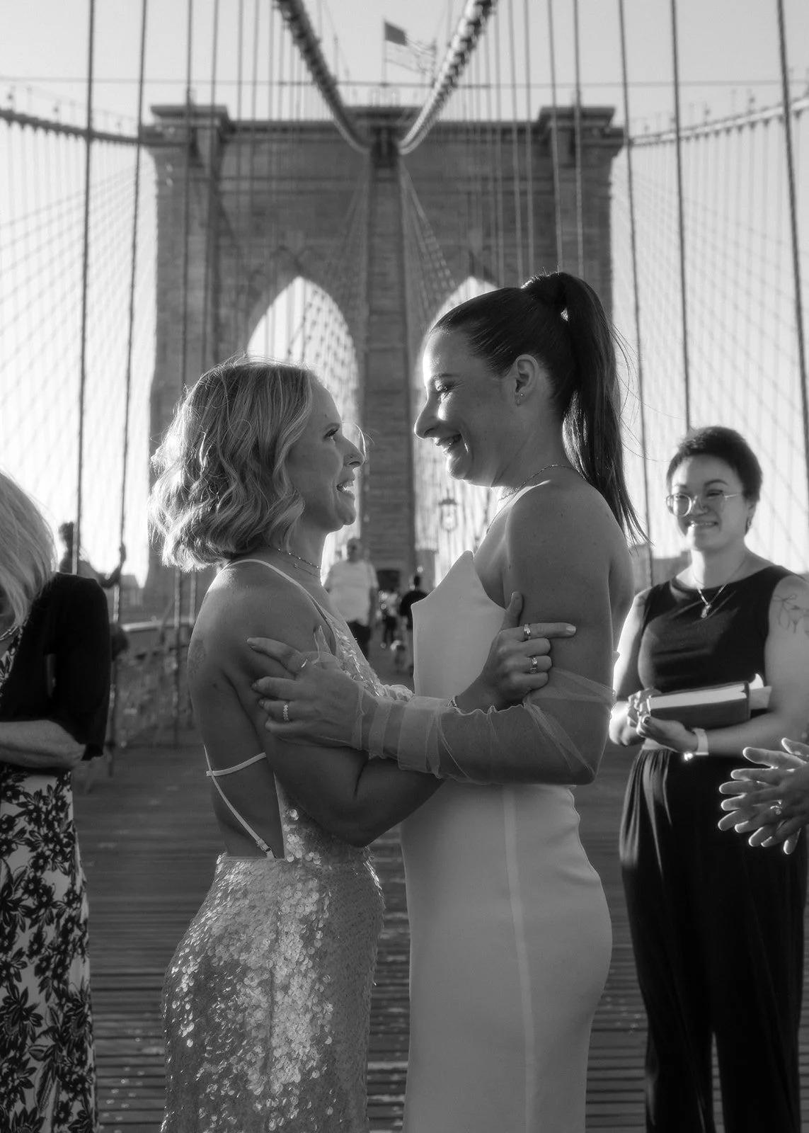LGBT brides embracing each other at their Cakewalk elopement on the Brooklyn Bridge at sunrise surrounded by their group of guests.