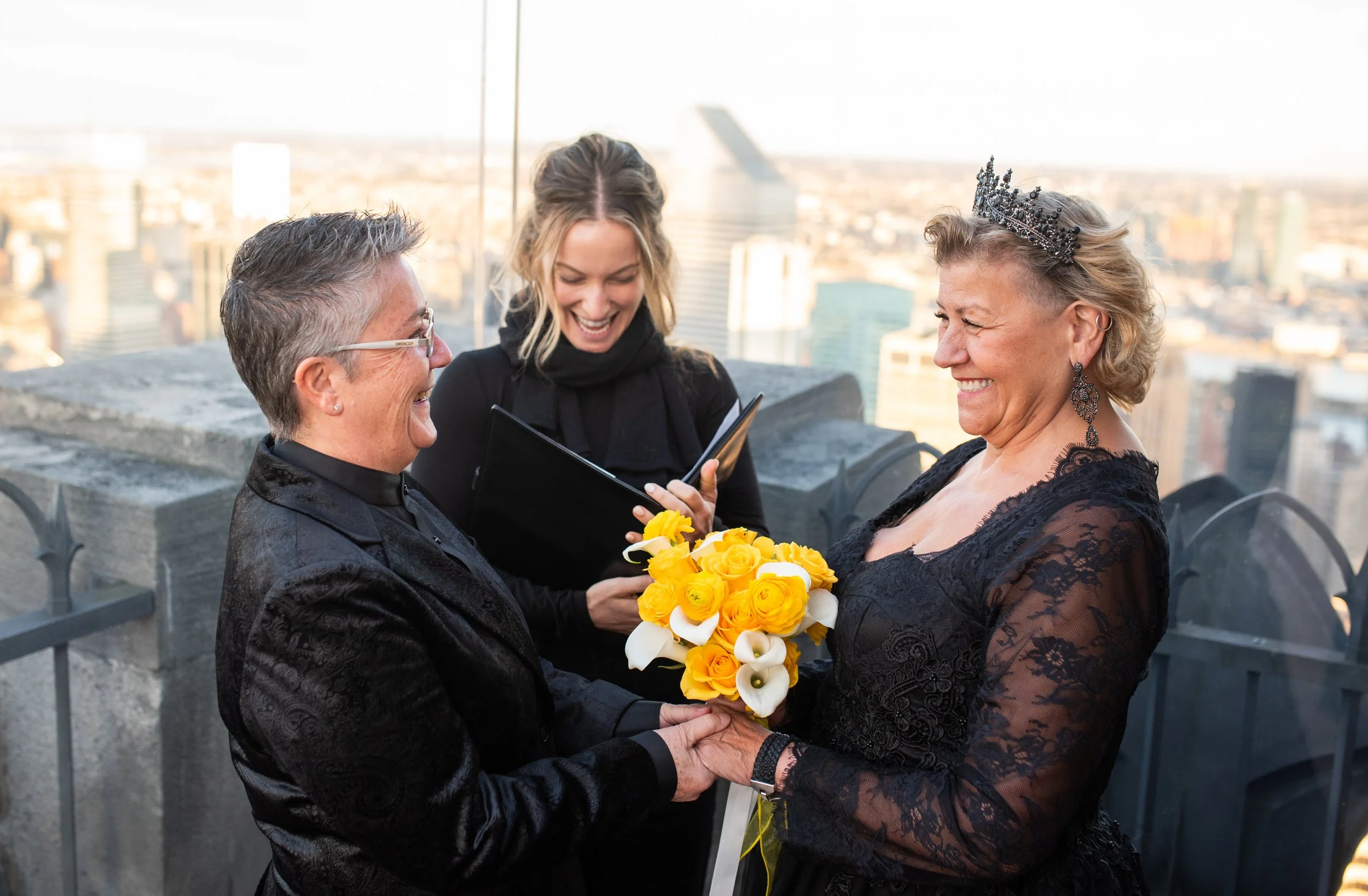 Two women wearing black and holding a yellow bouquet face each other smiling at the Top of the Rock as their LGBTQ Cakewalk officiant marries them.