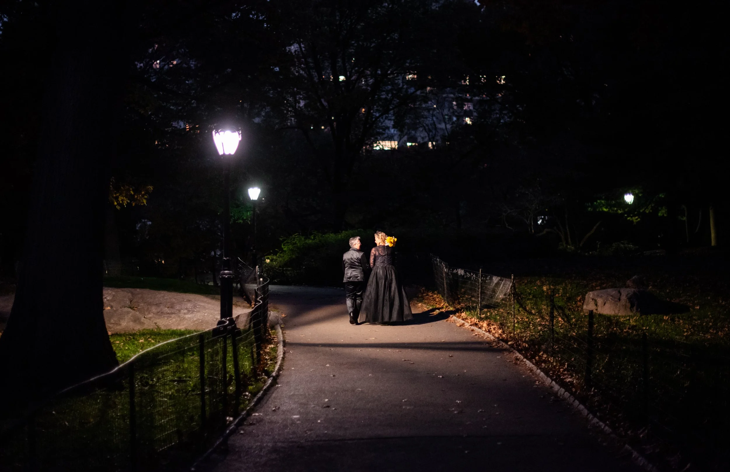 Two brides walking down a paved path in Central Park at night after their LGBTQ-friendly Cakewalk elopement.