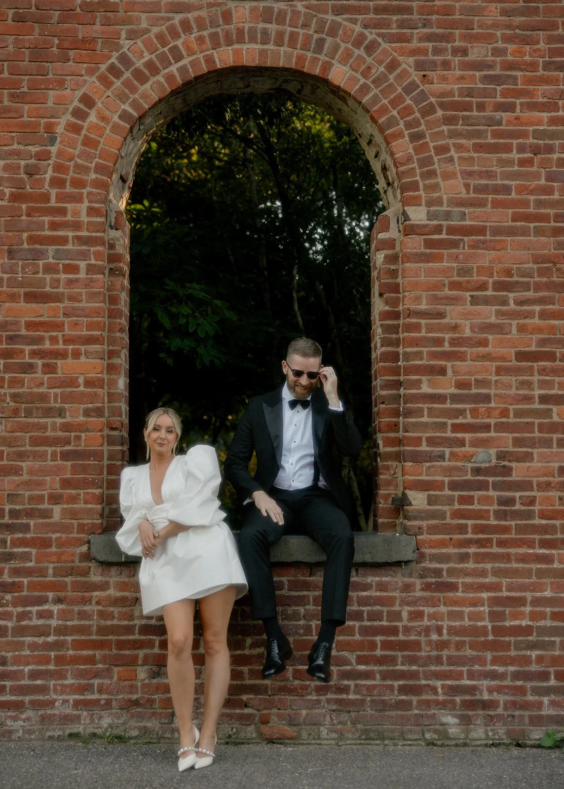 Bride and groom posing beneath a brick arch at an outdoor NYC elopement, with the groom sitting casually on the ledge in a tuxedo and the bride leaning against the wall in a short white dress, photographed in soft, cinematic light.
