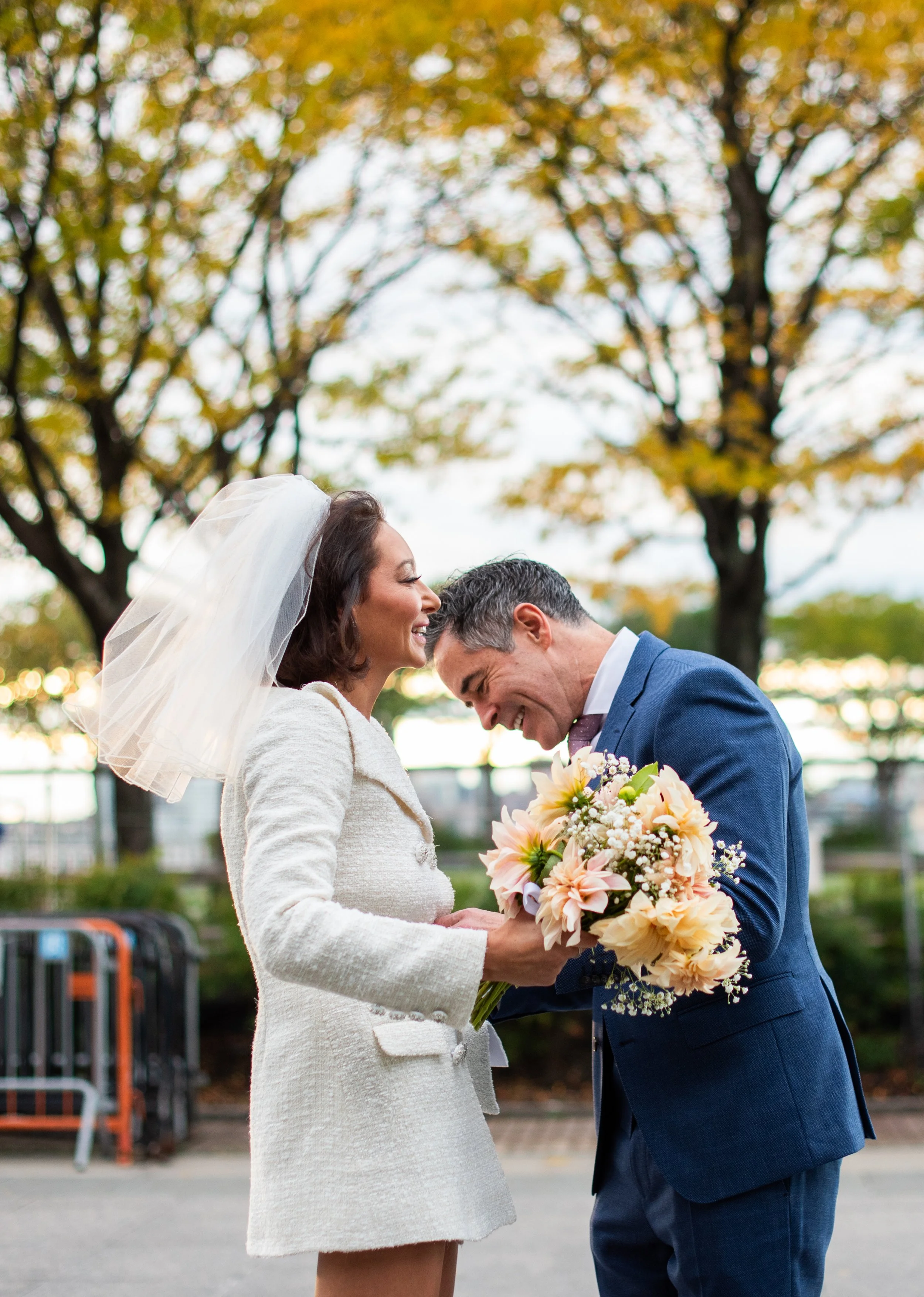 A woman in a white coat and a white wedding veil stands next to a man in a blue suit, both smiling as the man bends slightly to touch foreheads. The woman holds a bouquet of flowers. There are trees with autumn leaves in the background.