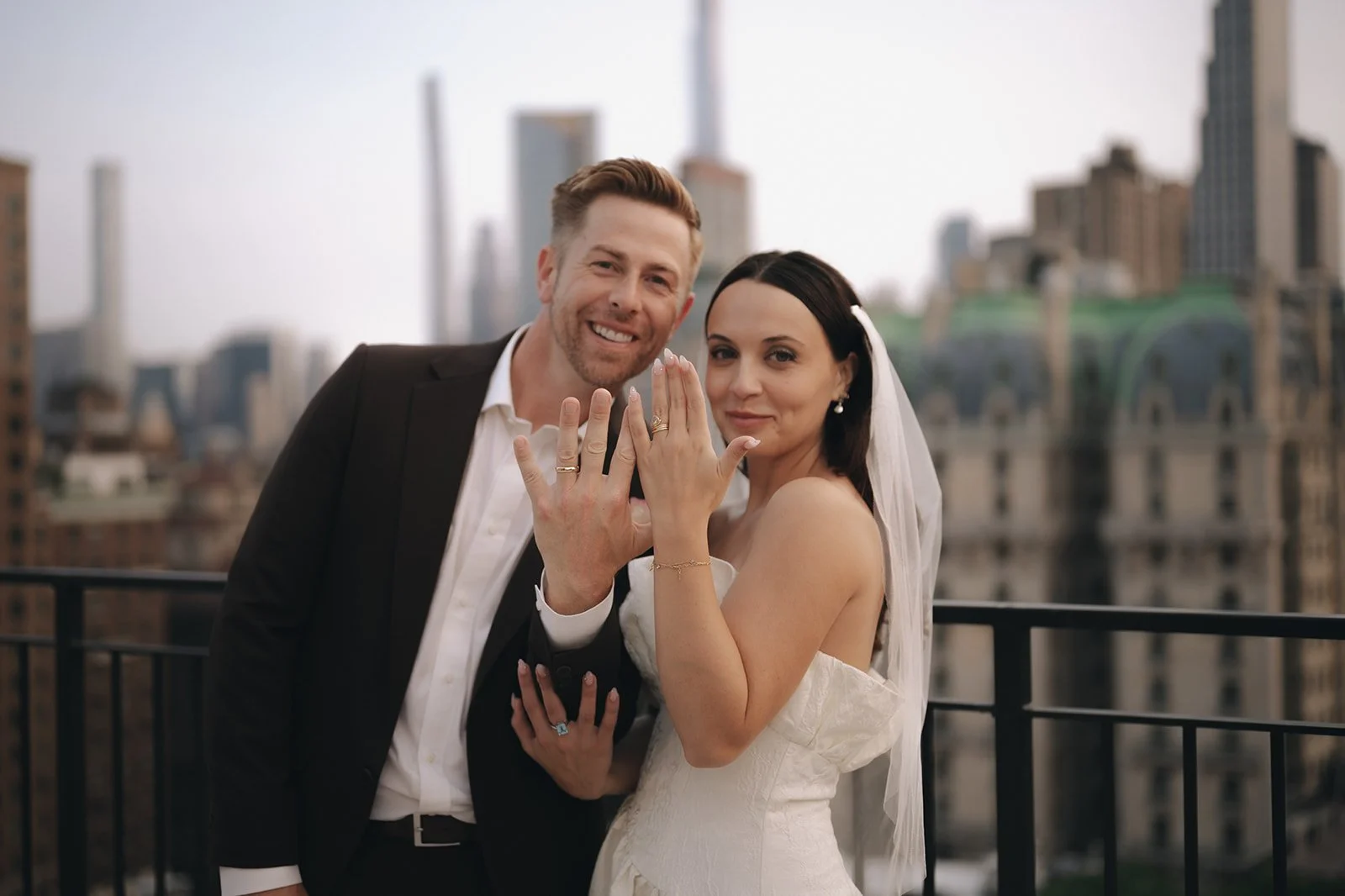A newlywed couple, a man in a black suit and a woman in a white wedding dress, showing their wedding rings on their fingers, with a city skyline in the background.