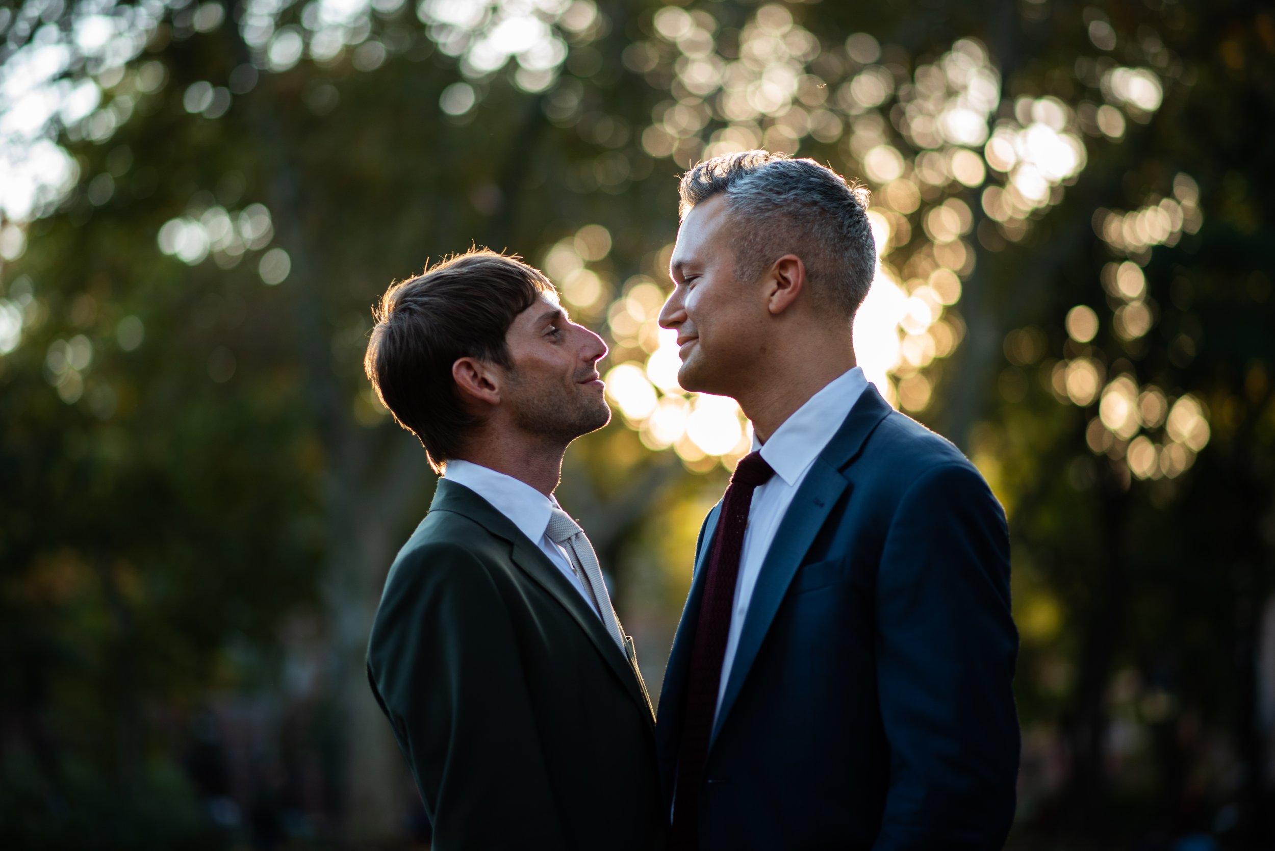 Two men looking into each other's eyes in Washington Square Park surrounded by trees and sunlight.