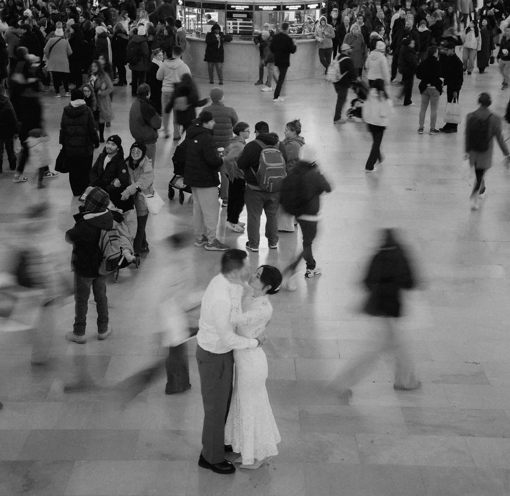 A crowded indoor space with many people walking around and some standing in groups. In the center, a couple is dancing closely together. The scene is lively and busy.