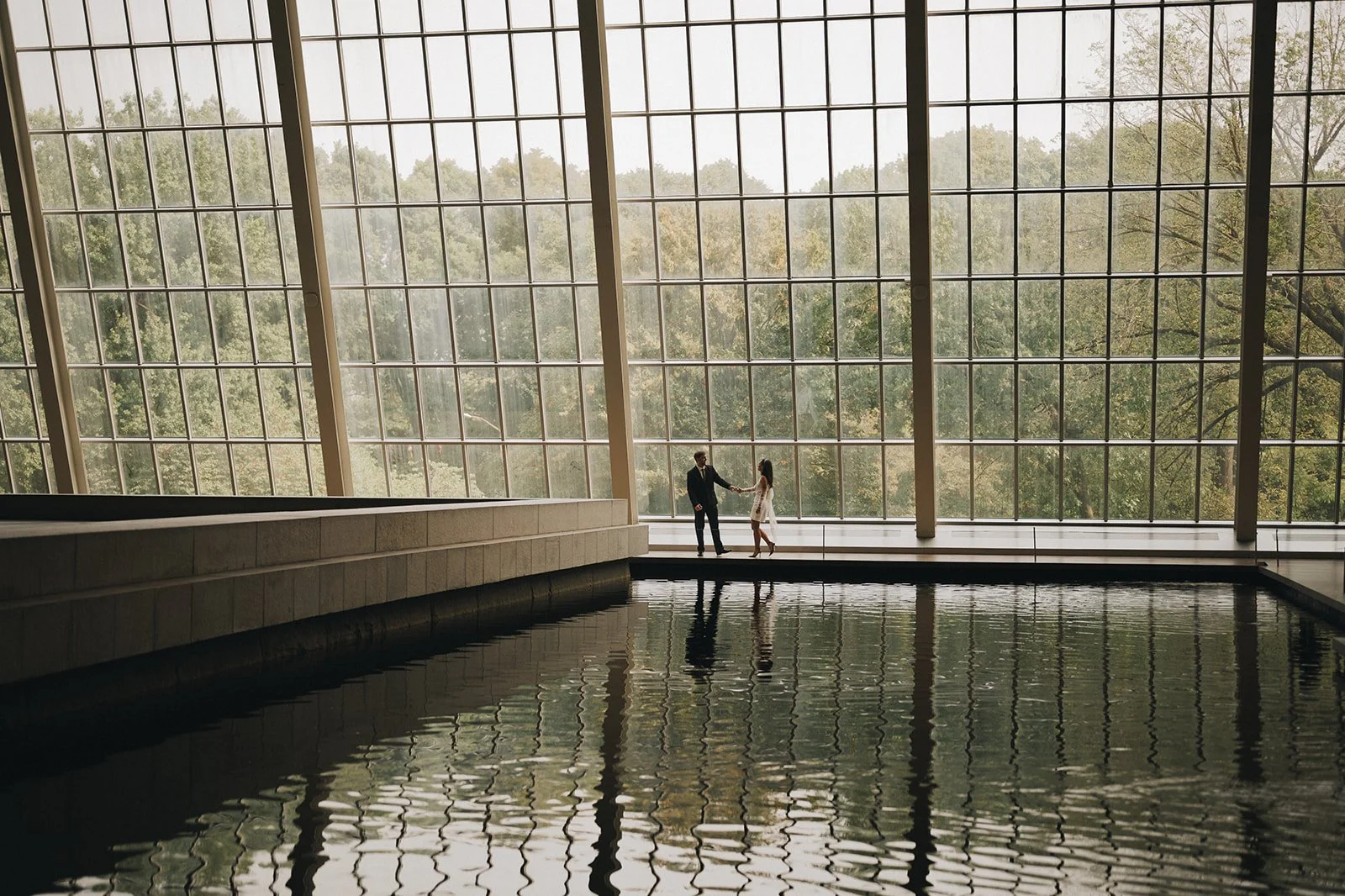 Bride and groom walking inside the Met Museum of Art inside a beautiful glass wall across from a reflecting pool of water.