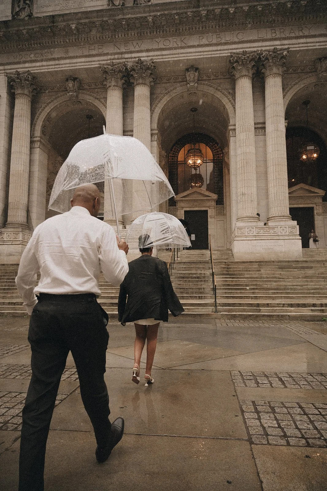 Bride and groom holding clear umbrellas, walking toward the the New York Public Library after their Cakewalk wedding ceremony.