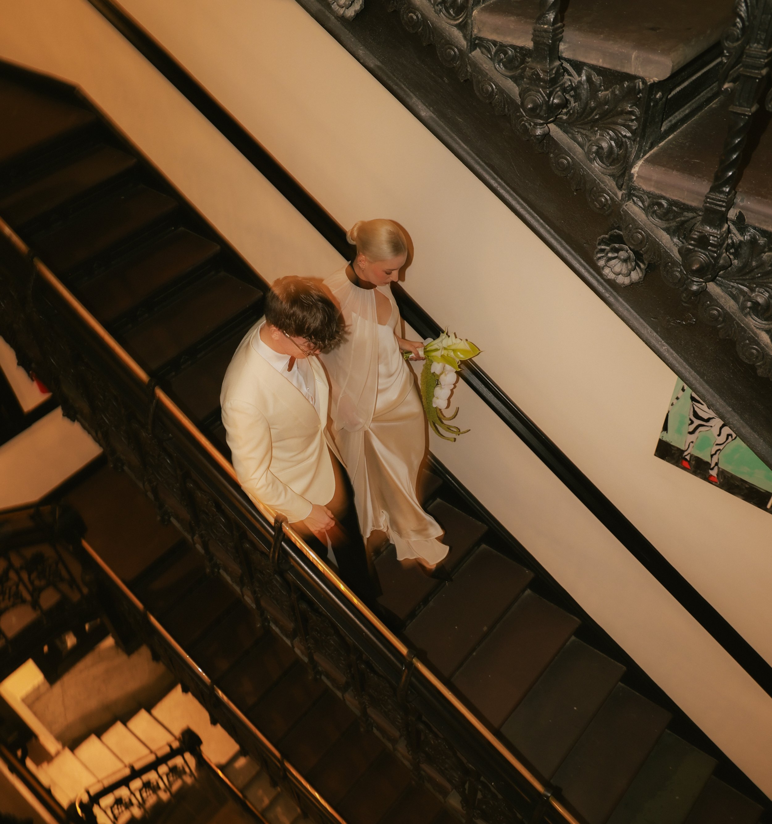 Bride, holding flowers, and groom walk down the stairs inside the Hotel Chelsea in Manhattan on the way to their their Cakewalk elopement.