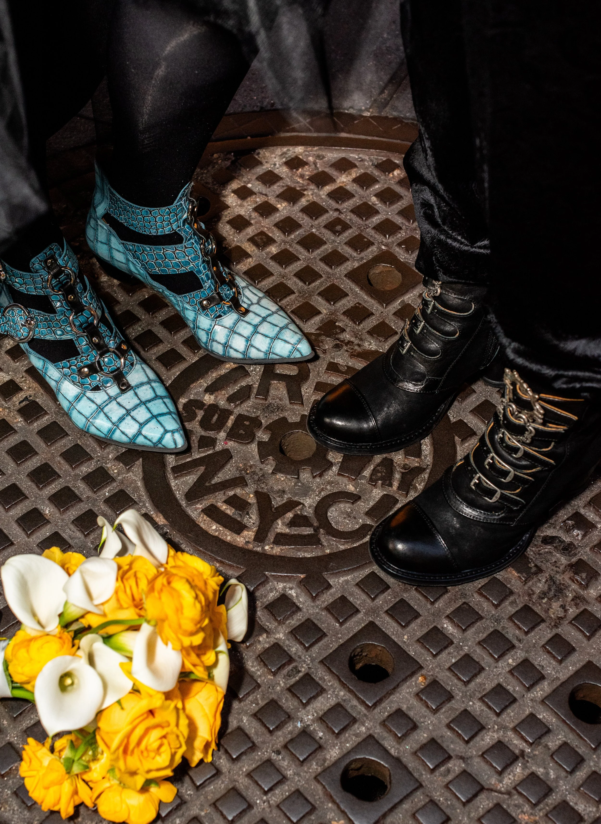 Close up of two pairs of boots standing on top of the NYC subway manhole with yellow and white flowers.