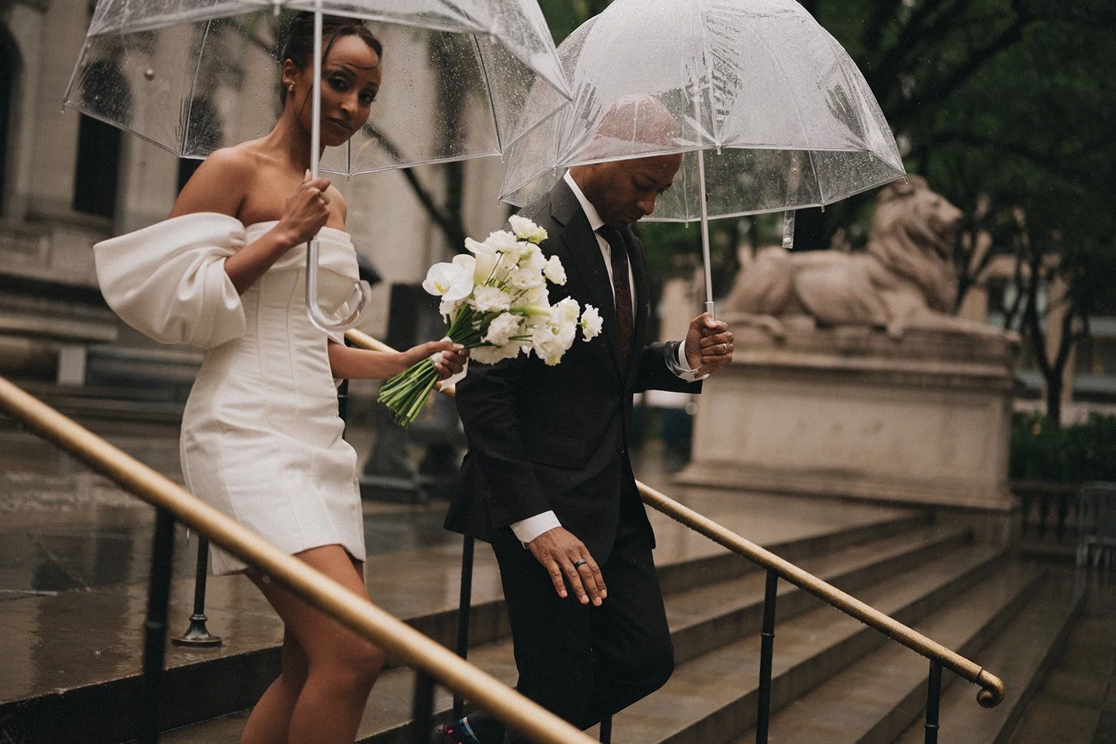 Bride and groom holding clear umbrellas, walking down stairs at the New York Public Library after their Cakewalk wedding ceremony.