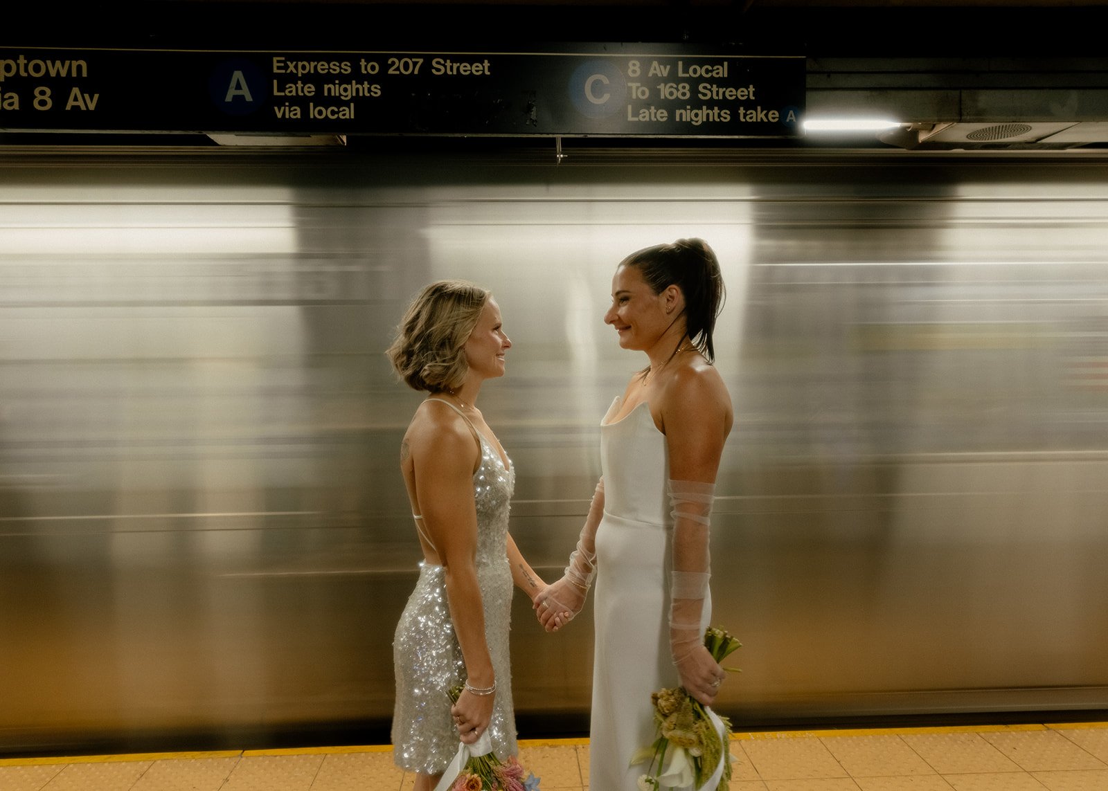 Australian lesbian brides standing in a New York City subway station with a moving train in the background following their Cakewalk elopement ceremony.