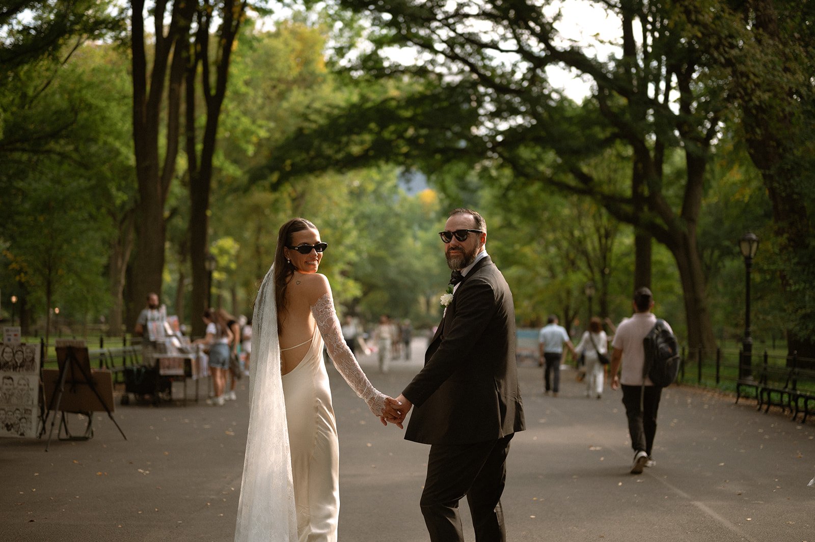 Bride and groom wearing sunglasses, looking back at camera while walking down a tree-lined path in Central Park after their Cakewalk micro wedding.
