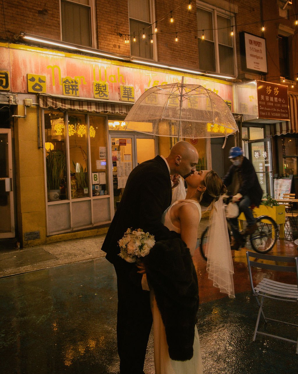 A couple sharing a romantic kiss under a clear umbrella on a rainy night in Chinatown, NYC, standing outside Nom Wah Tea Parlor during their elopement.