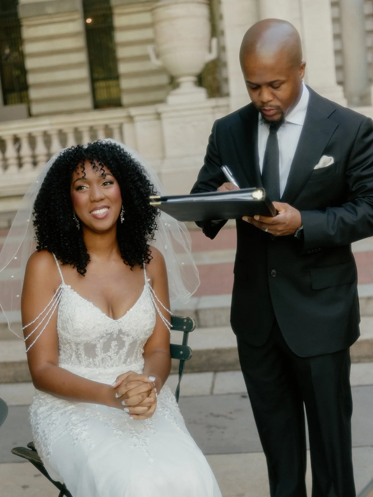 Bride sitting and groom standing while signing marriage license paperwork after being married by their Cakewalk elopement ceremony at Bryant Park.