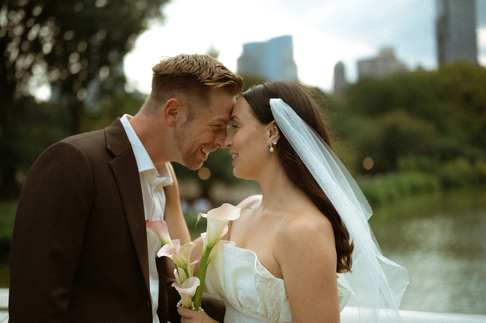 A newlywed couple touches foreheads while smiling, outdoors with a blurred cityscape and trees in the background. The bride wears a white wedding gown and veil, holding a bouquet of calla lilies. The groom wears a brown suit and white shirt.