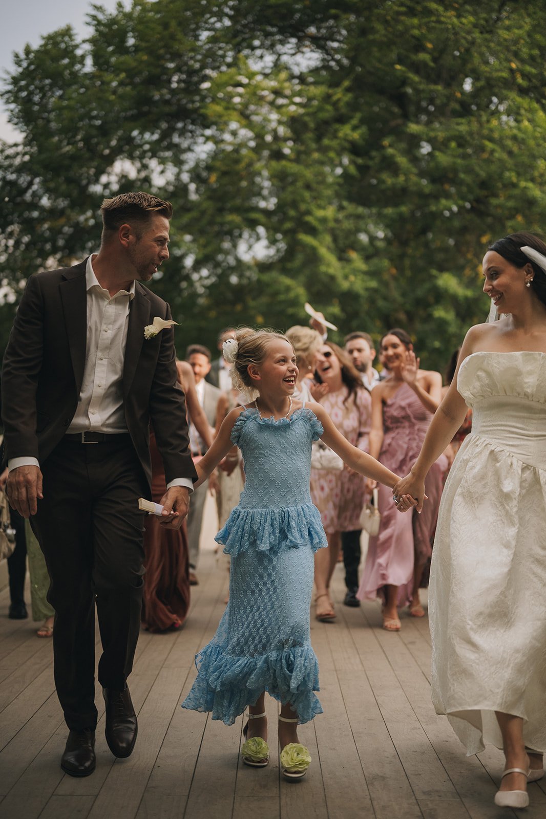 Bride, groom, and daughter walking hand-in-hand with family following behind across Bow Bridge after their Central Park elopement ceremony by Cakewalk.