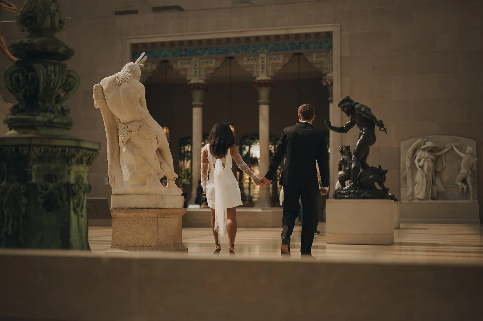 Bride and groom walking inside the Met Museum of Art among the sculptures.