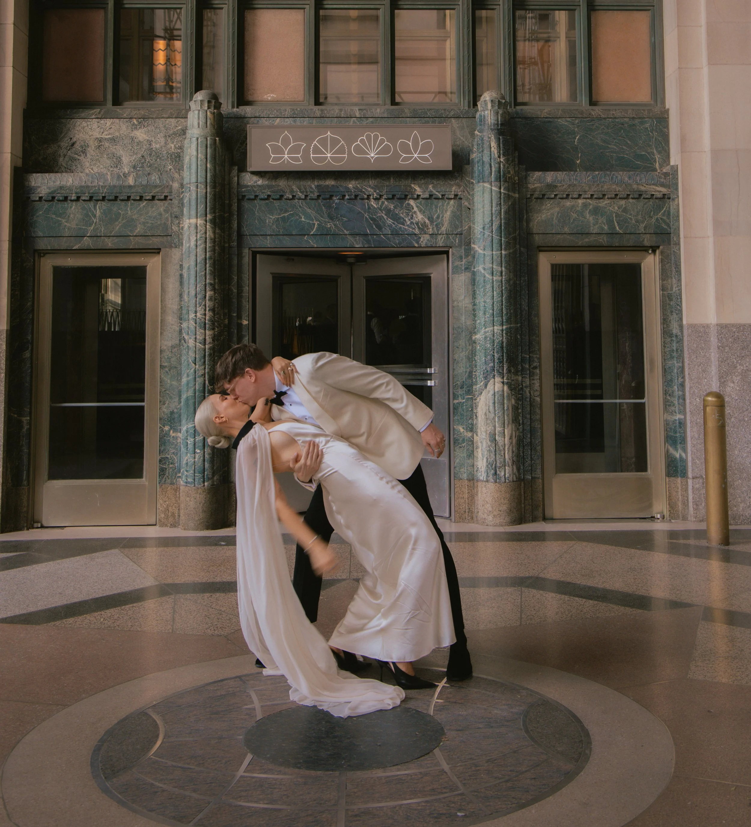 Bride and groom kissing in a plaza with old art deco architecture behind them in New York City.