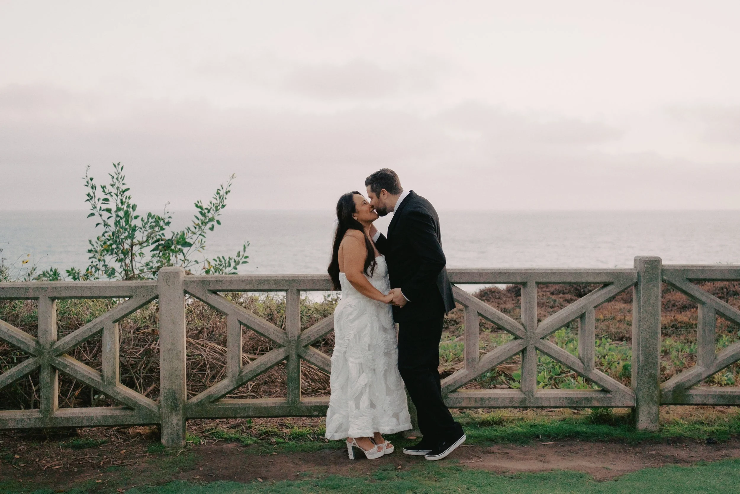 Bride and groom kissing on cliffside in Santa Monica with a pretty fence and the ocean behind them.