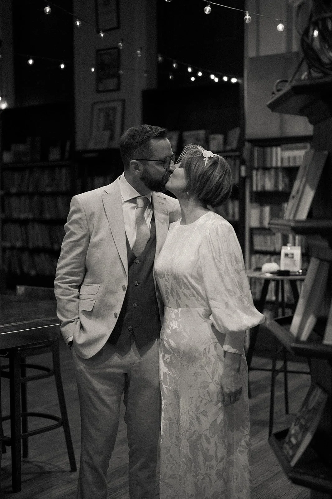 Bride and groom kissing in Housing Works Bookstore with books behind them and string lights overhead during their Cakewalk elopement.