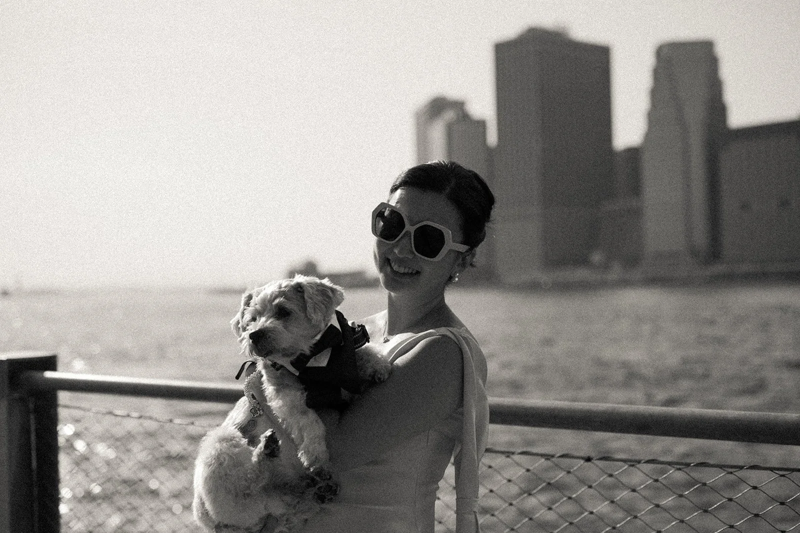 Couple holds their dog during a Brooklyn waterfront elopement, with the NYC skyline in the background, photographed in a documentary style.