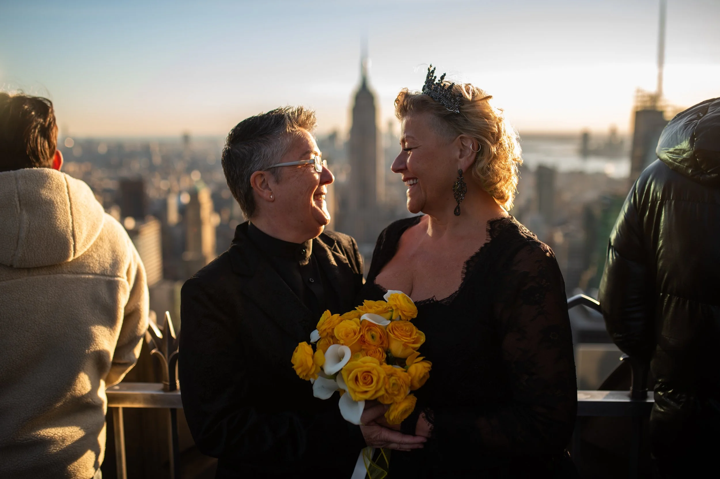 Two women wearing black and holding a yellow bouquet face each other smiling at the Top of the Rock with the Empire State Building in the background.