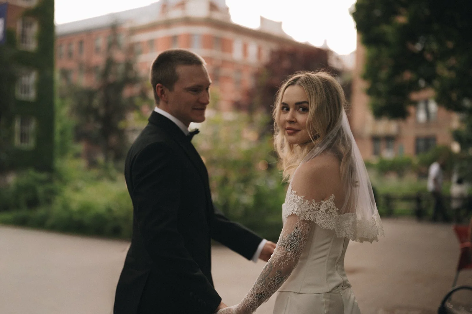 A bride and groom holding hands outdoors in a park, with trees and buildings in the background during sunset.