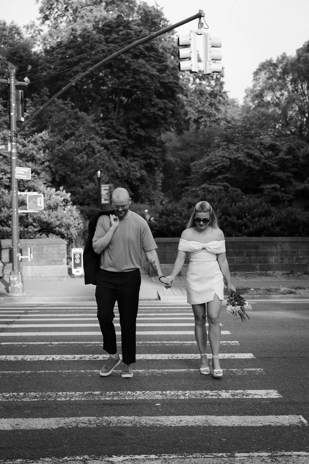 A black and white photo of a couple holding hands and walking across a crosswalk. The woman is wearing an off-shoulder dress and high heels, holding a bouquet of flowers, and the man is dressed casually with tattoos on his arm, carrying a jacket over