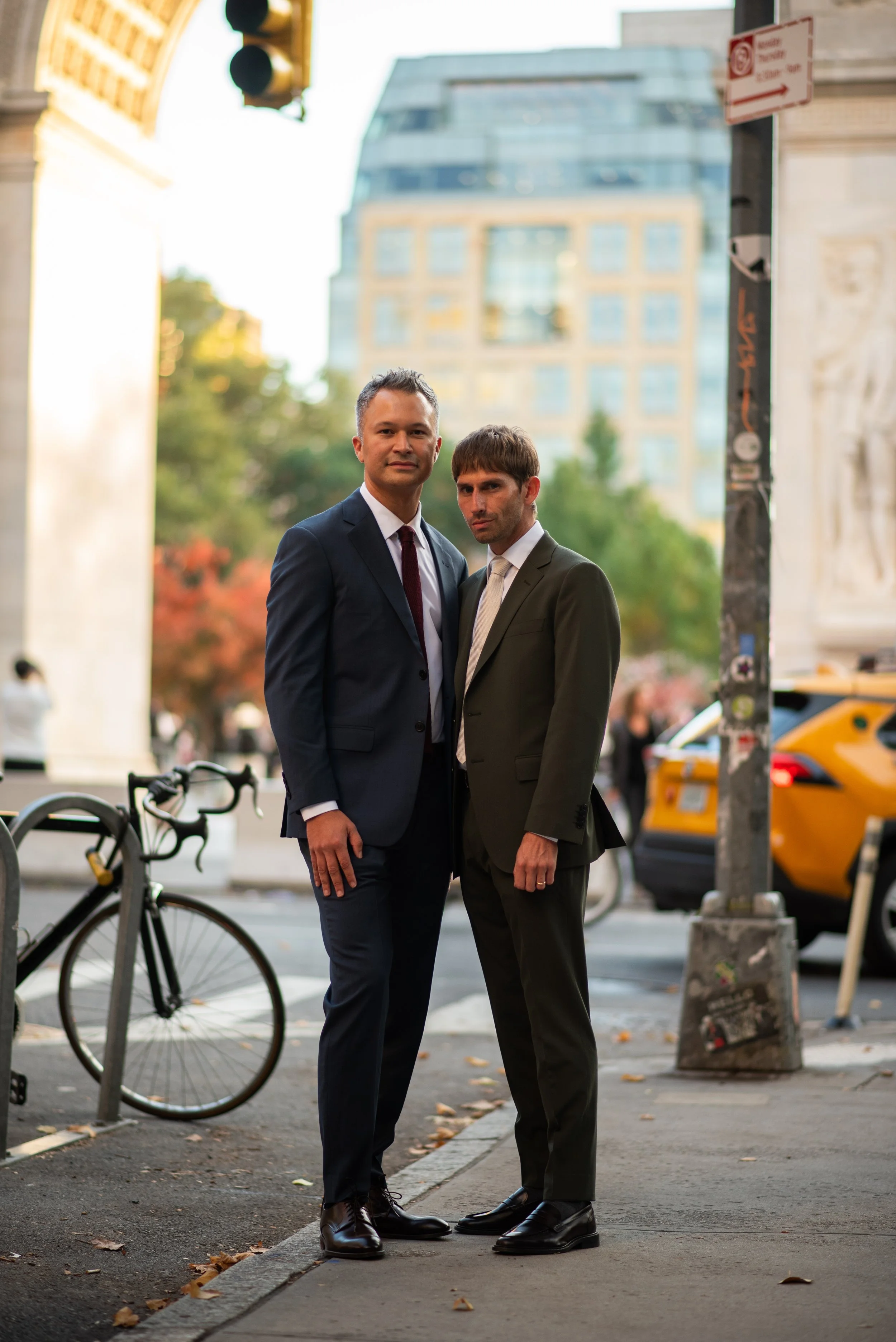 Two men standing on the sidewalk with a bike, yellow taxi, and the arch at Washington Square Park behind them.