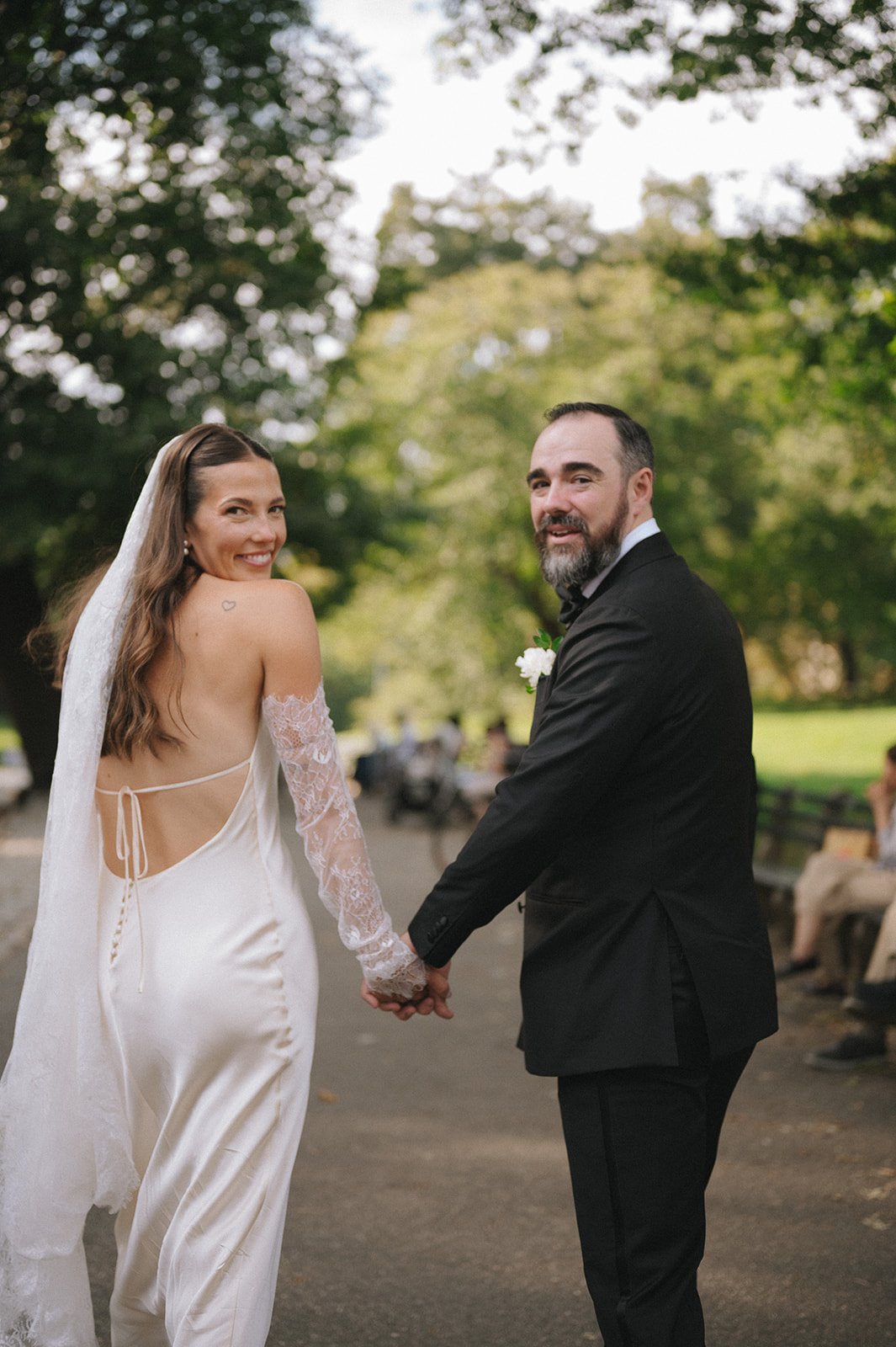 Bride and groom walking to their wedding ceremony in Central Park, looking back over their shoulders at the camera.