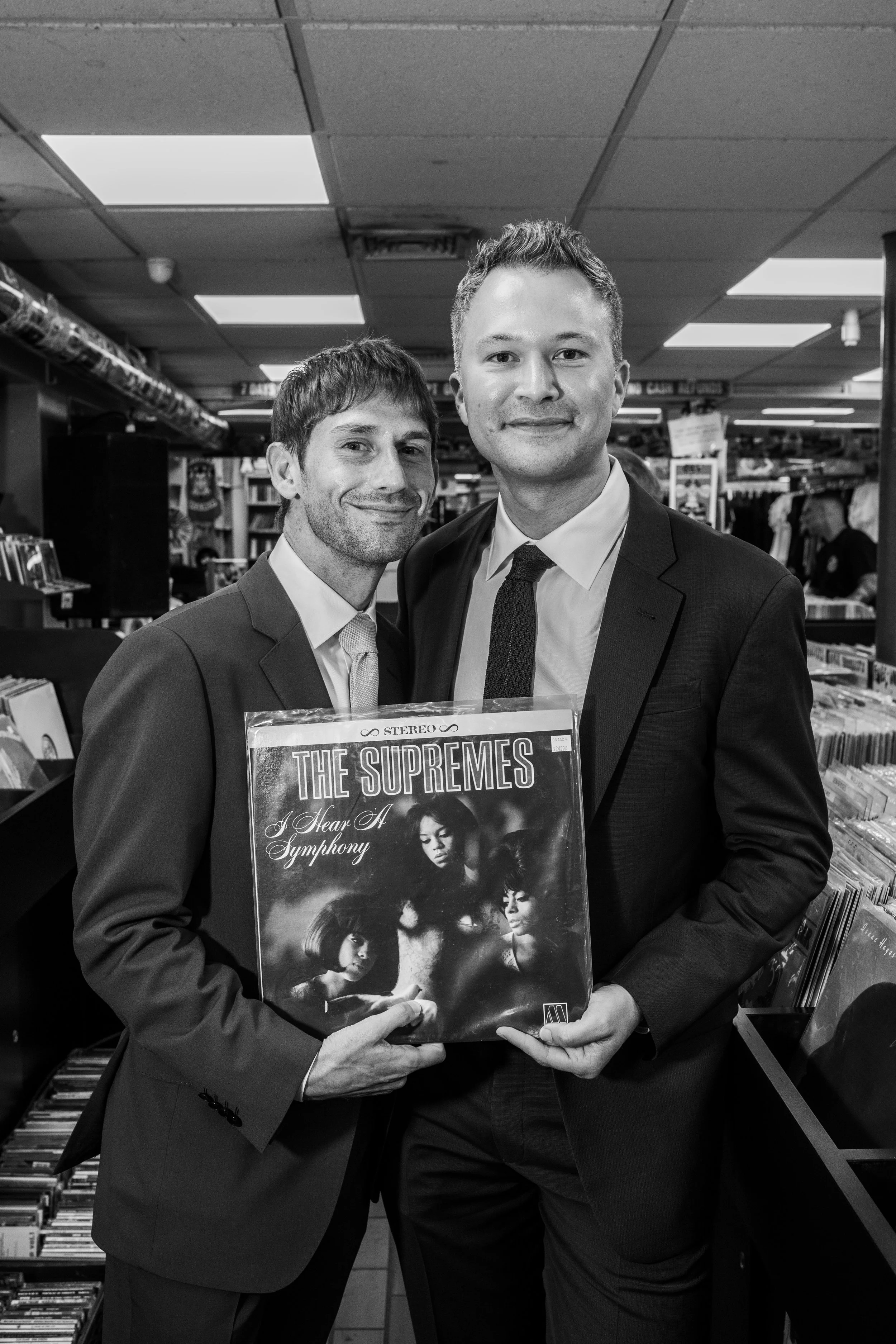 Two grooms in suits holding a Supremes album in a record store in black and white.