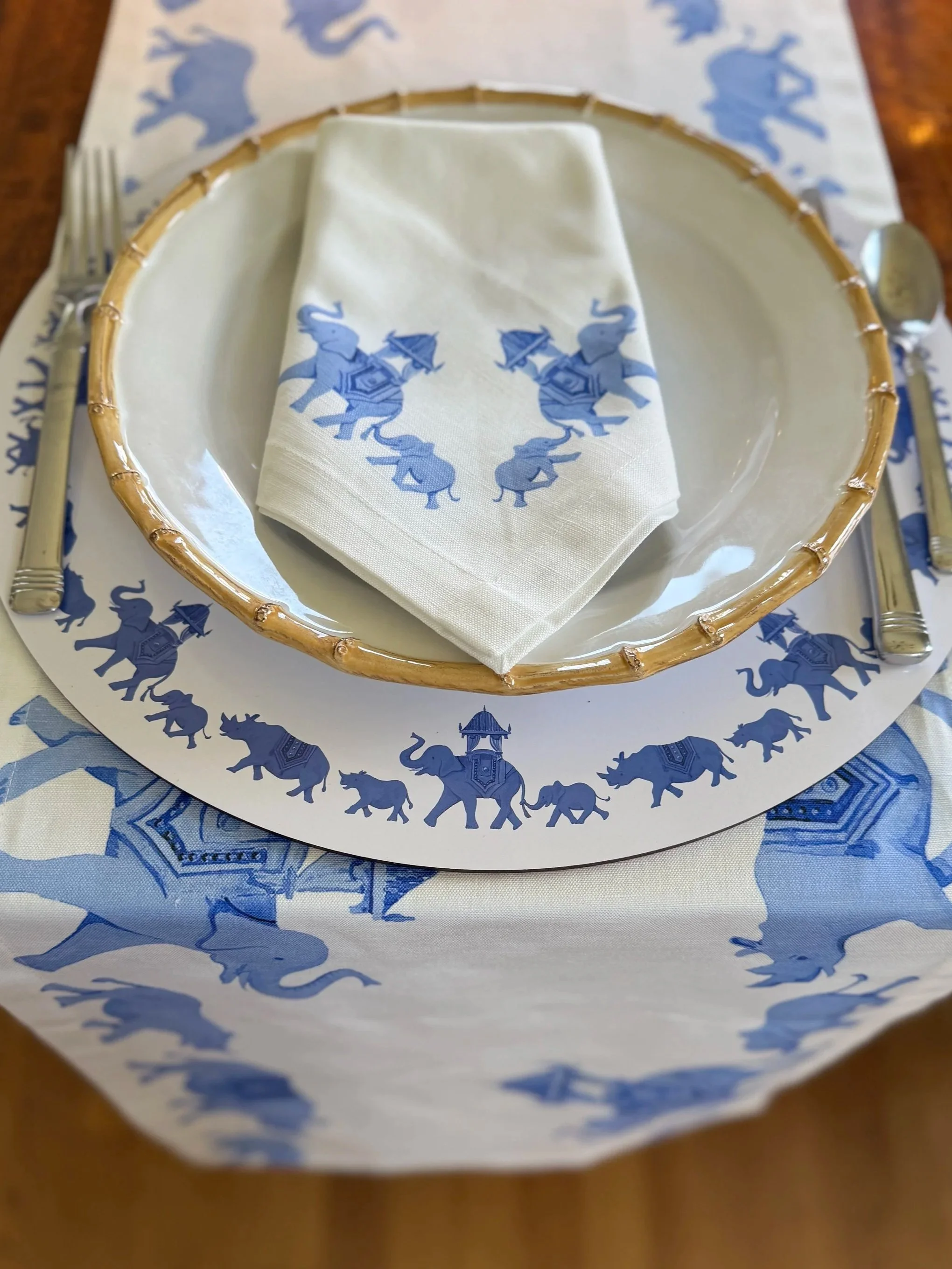 A dinner setting with a white napkin featuring blue elephants holding umbrellas, placed on a white plate with a bamboo-like rim. The plate is on a blue and white elephant patterned tablecloth and paper placemat, with a fork on the left and a spoon on the right.