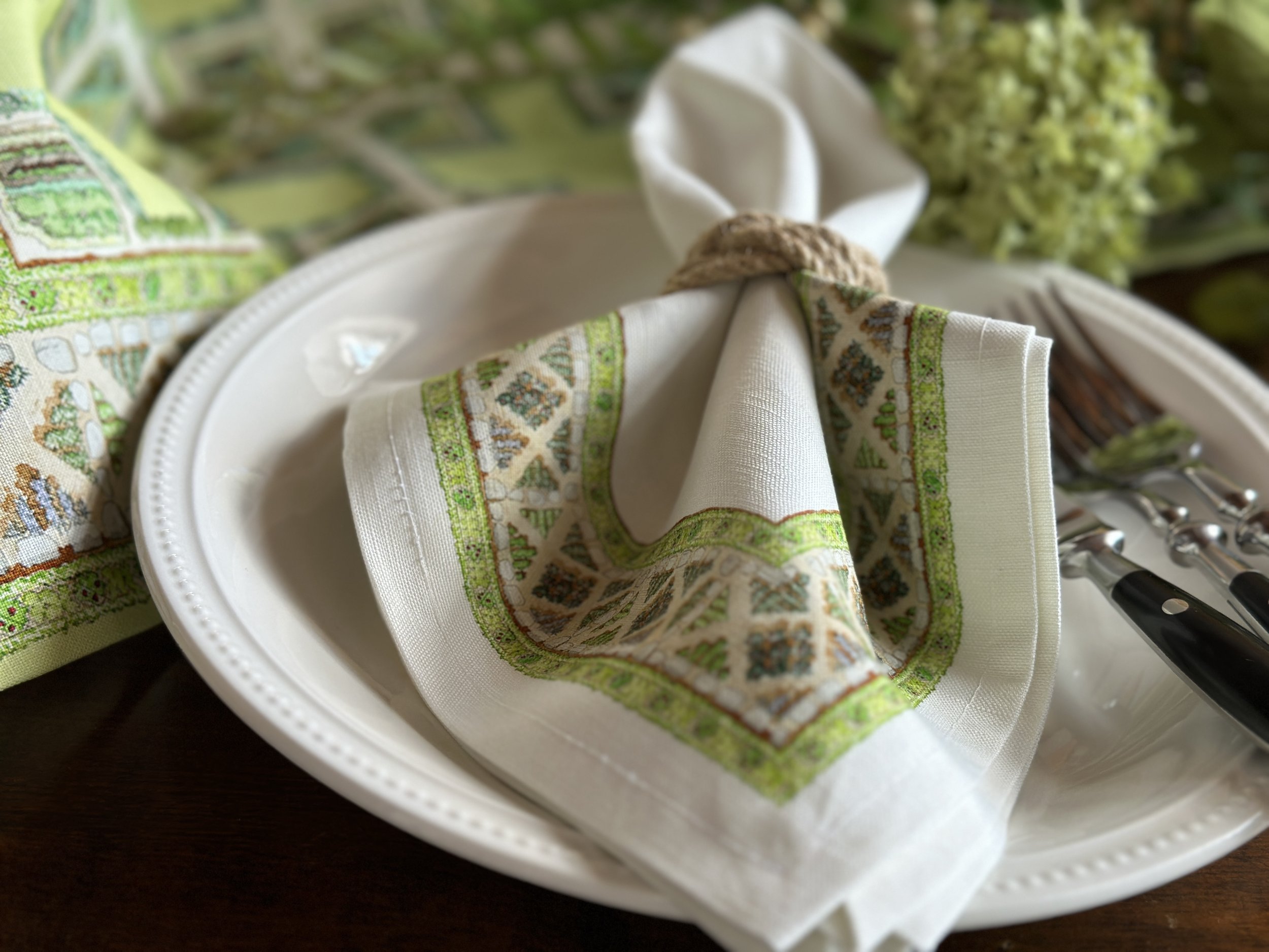 Close-up of a decorative napkin with a Christmas pattern tied with a jute string, placed on a white ceramic plate, with cutlery beside it, on a dark wooden surface.
