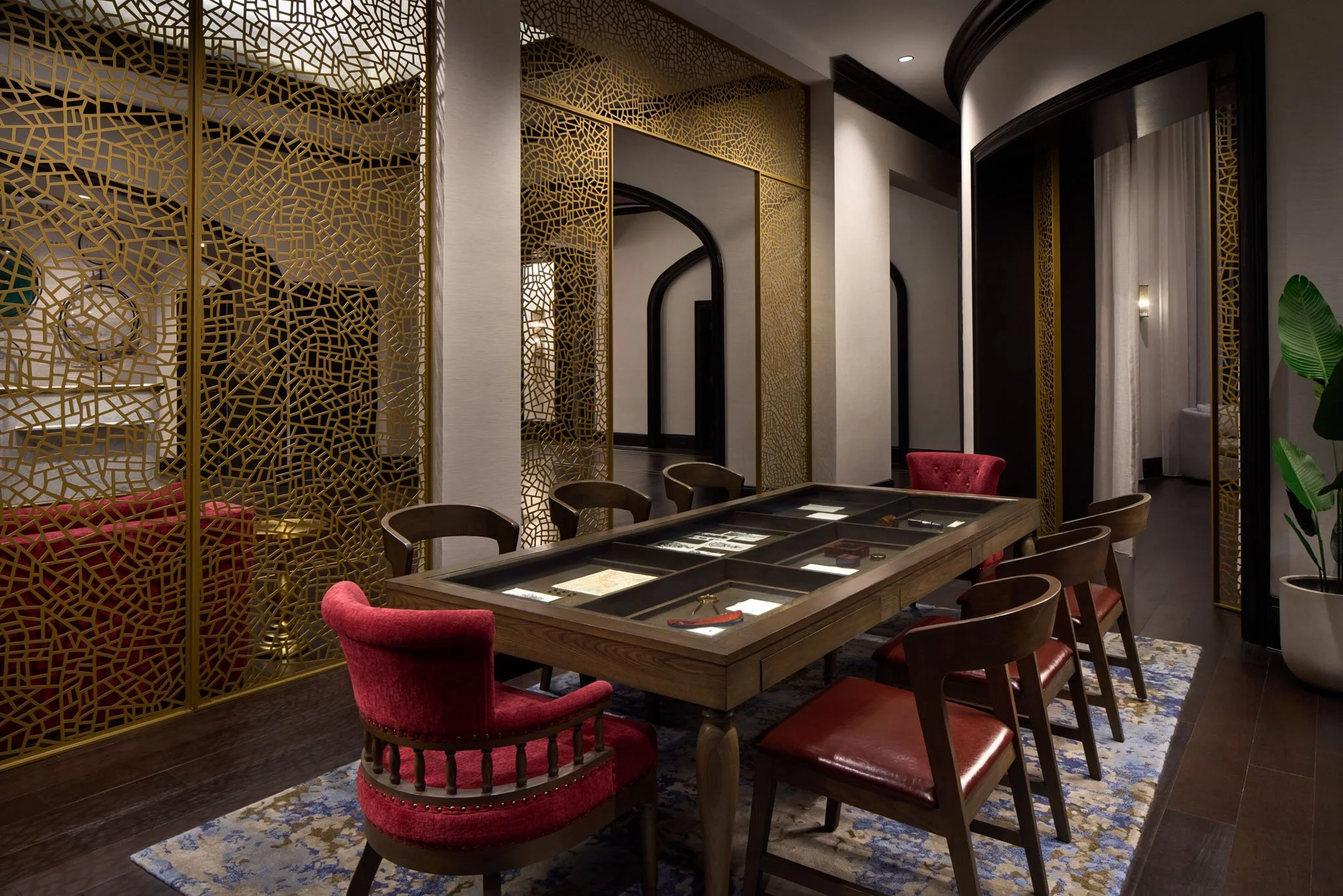 A stylish room with a long wooden table enclosed in a glass display case, surrounded by six chairs with red and brown upholstery. The room features intricate gold lattice screens, dark wood flooring, and a patterned rug. There are potted plants and a