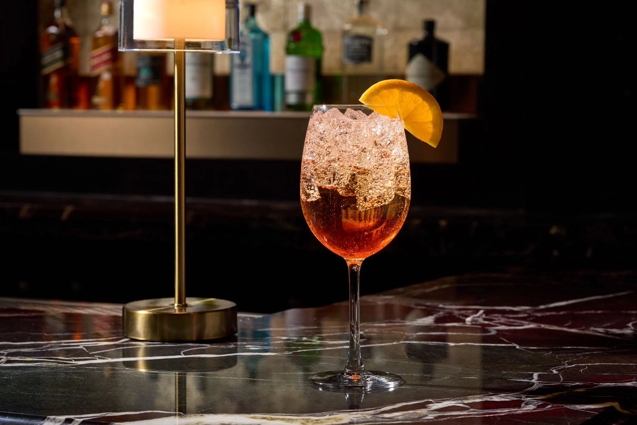 A cocktail with ice and an orange wedge garnish in a wine glass, placed on a marble countertop with background shelves of liquor bottles.