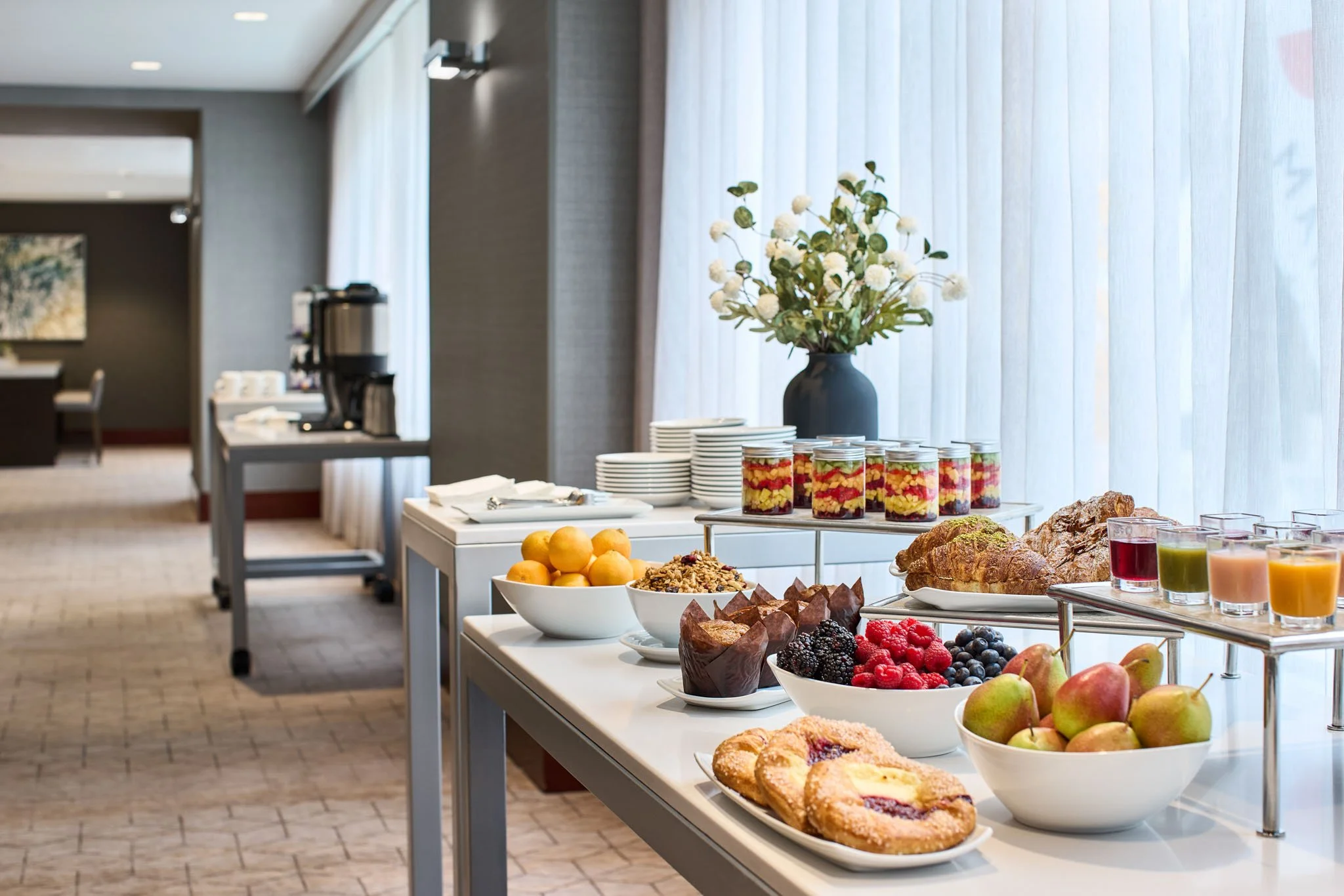 Breakfast buffet with bowls of fruit, pastries, yogurt jars, and juice glasses set on a table near a large window with sheer curtains in a hotel dining area.