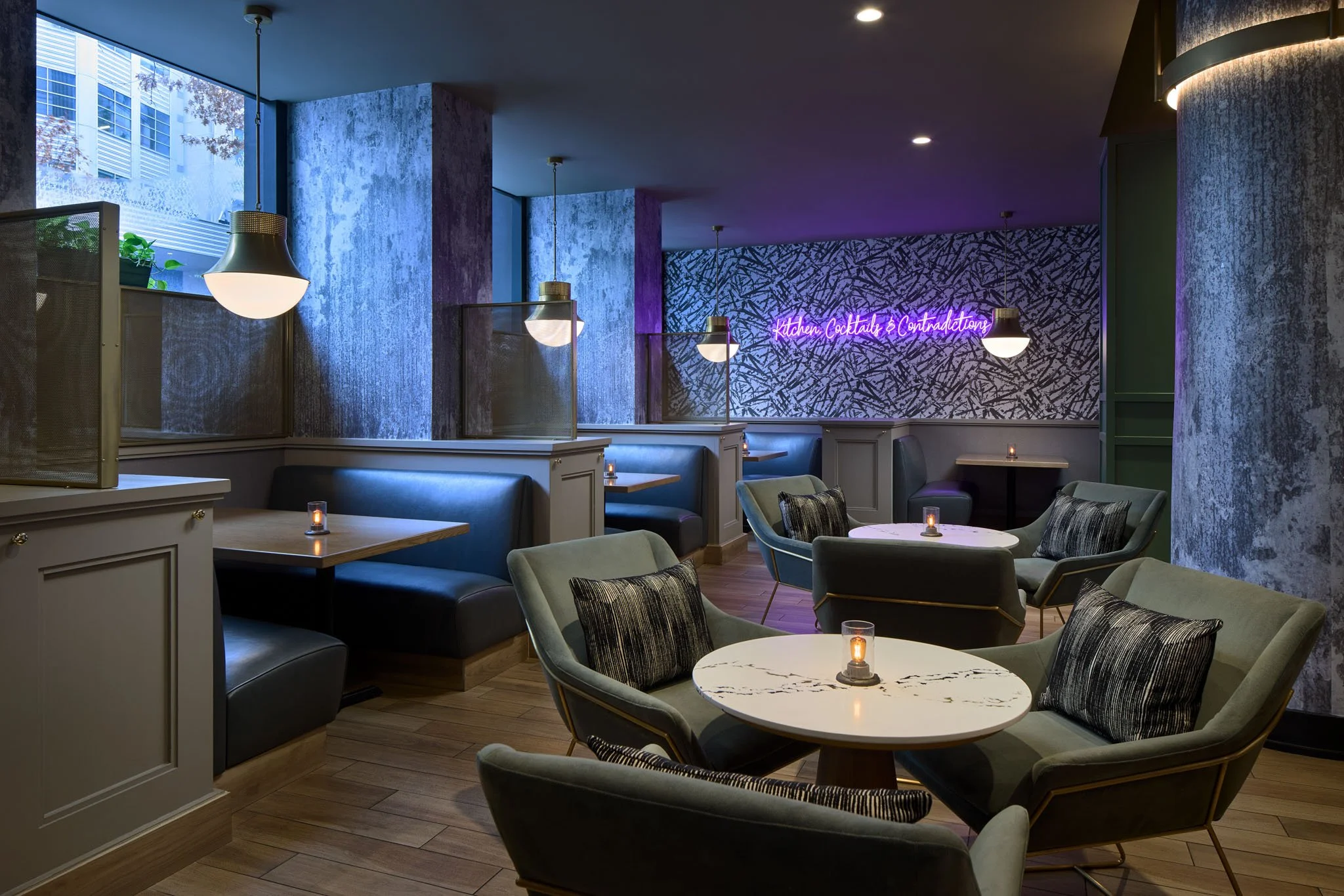 Interior of a modern restaurant or cafe with cozy seating, small lit candles on tables, large windows letting in natural light, textured walls, and a neon sign that reads 'Kitchen, Cocktails & Contradictions' in pink