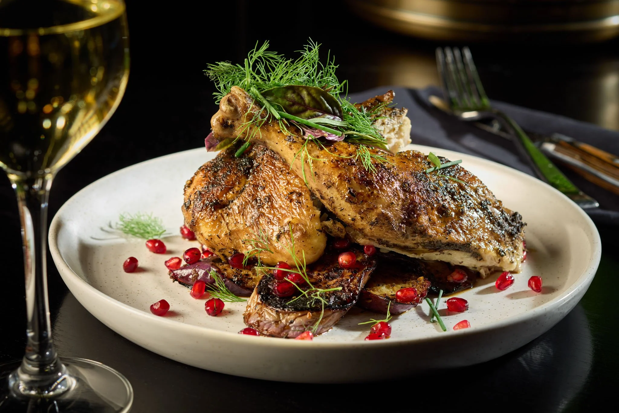 Grilled chicken served on a white plate with garnishes, pomegranate seeds, and herbs, with a glass of white wine nearby.