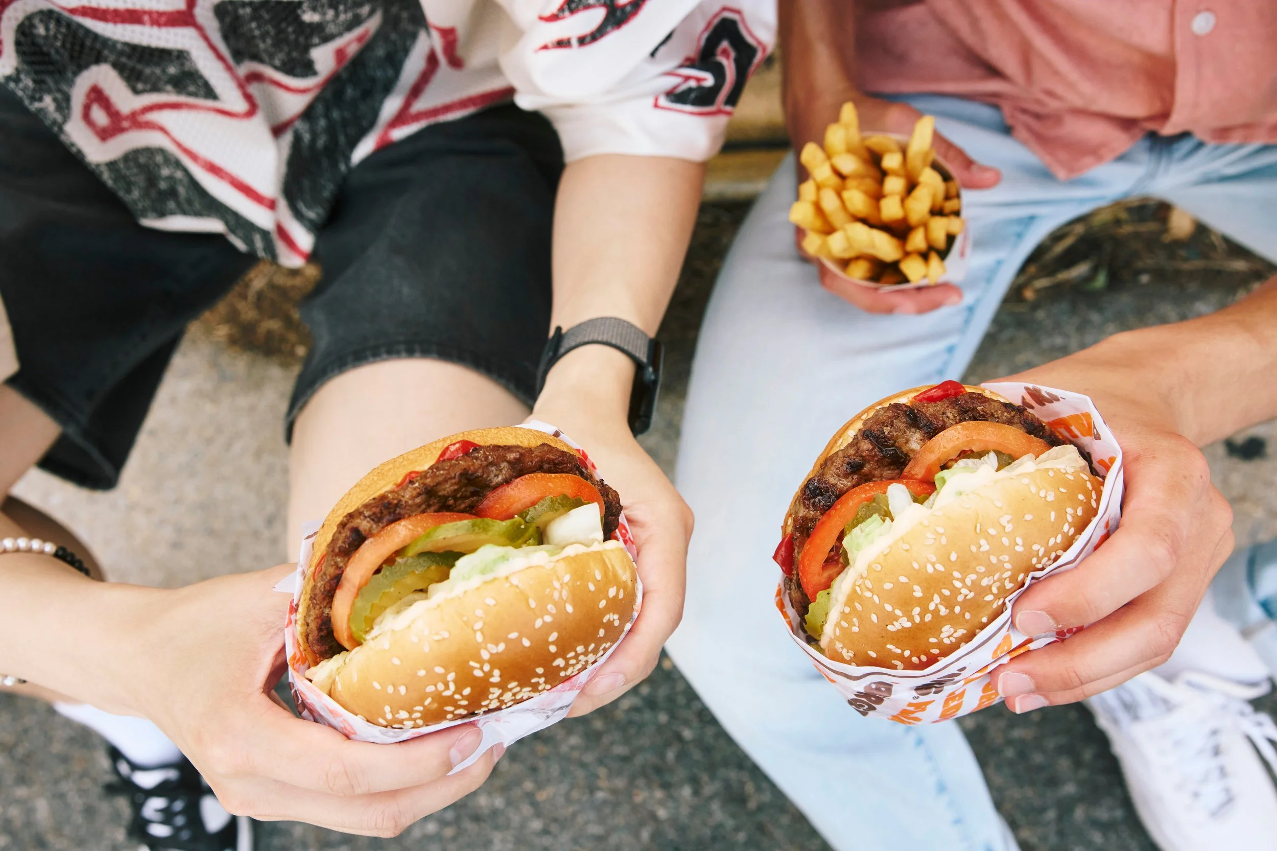 Two people holding Whoppers with lettuce, tomato, pickles, and beef patties, and two people holding cups of French fries, with one person wearing a black and white striped shirt and black shorts, and another wearing a pink shirt and light blue pants.