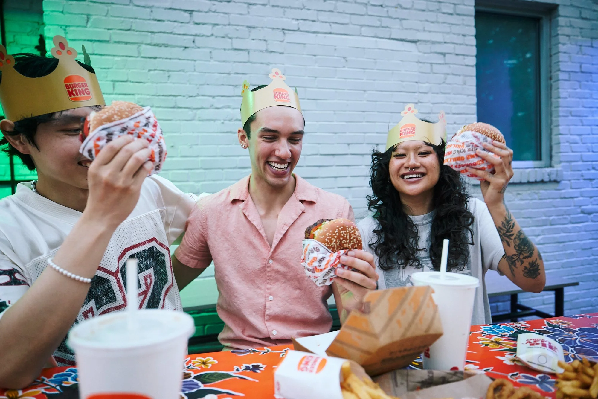 Three young people at a Burger King-themed birthday party, wearing themed paper crowns, holding Whopper sandwiches, and smiling. They are seated at a table with colorful food packaging and drinks, against a background of a brick wall with blue and gr